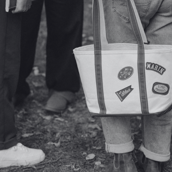 Close-up of a person holding a large striped tote bag with patches, standing on dirt ground, with other people's legs visible in the background.