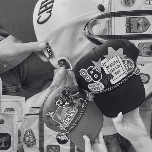 Two baseball caps with patches and chains being held up, one black and one light-colored, with various patches and pins, against a background of clothing or accessories with patches.