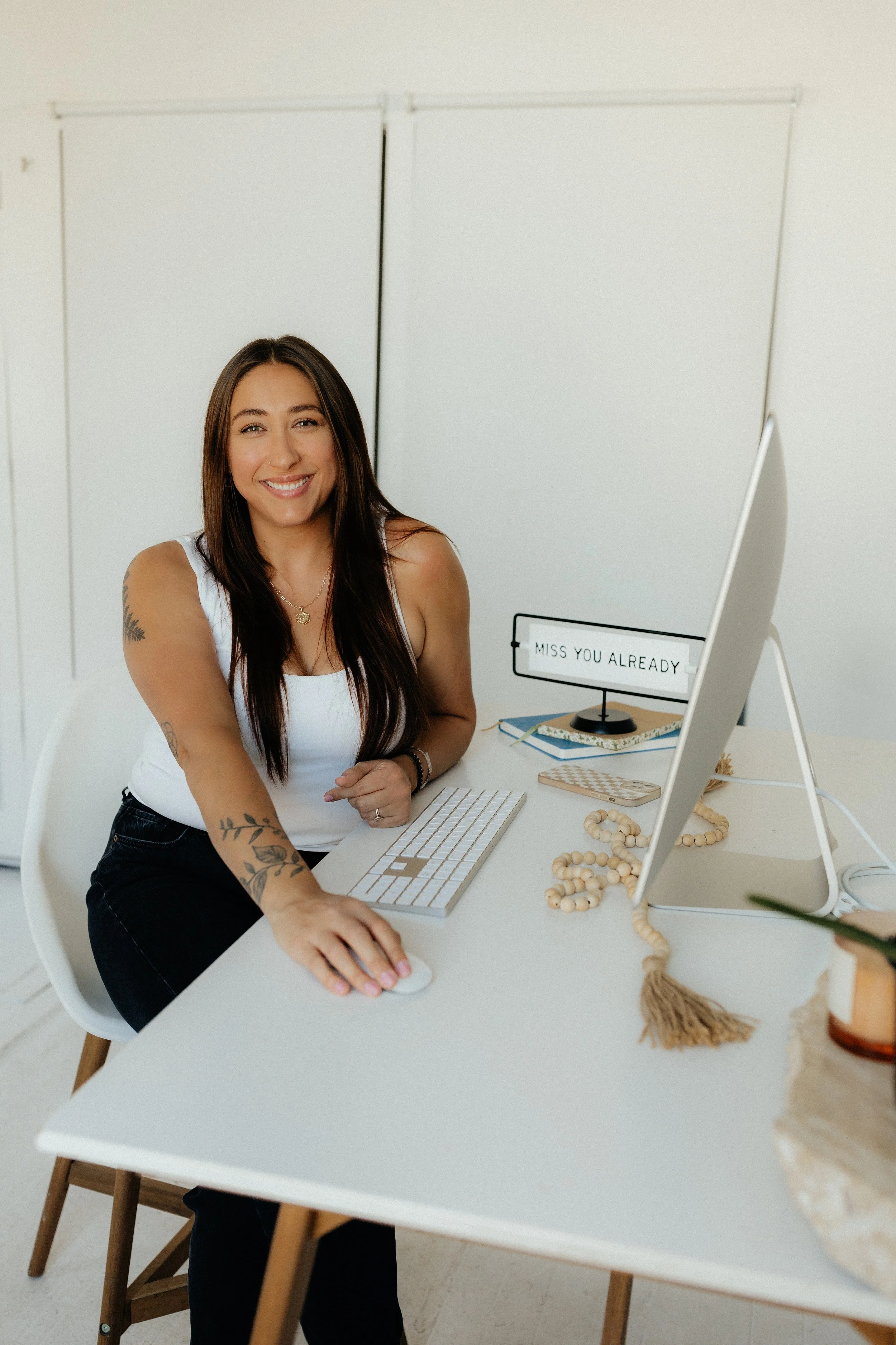 A smiling woman with long dark hair sitting at a white desk, using a computer mouse and smiling at the camera. The desk has a keyboard, a monitor, a sign that reads 'Miss You Already,' books, a beaded necklace, and a small decorative tassel.