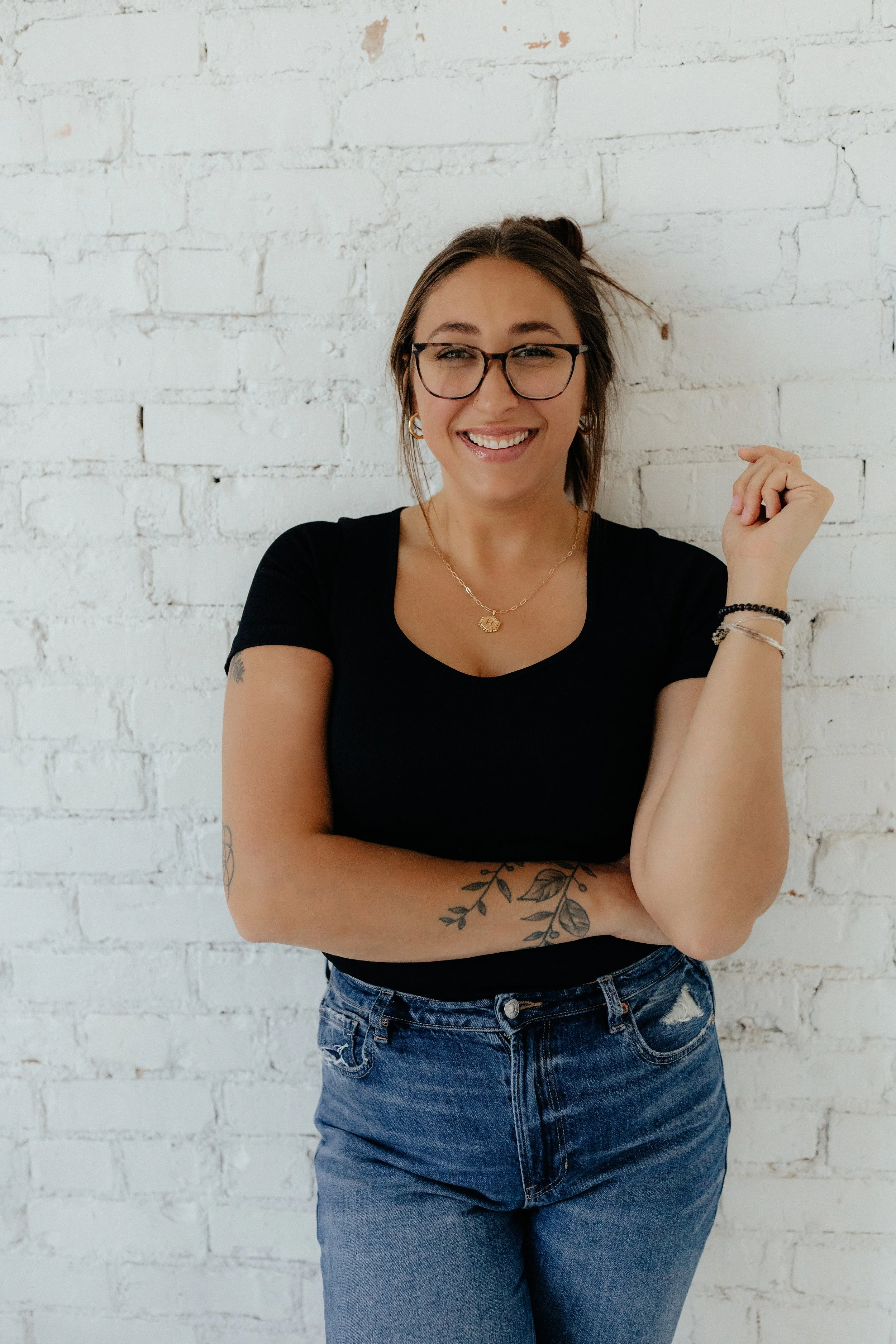 A young woman with glasses and tattoos smiling, standing against a white brick wall, wearing a black t-shirt and jeans.