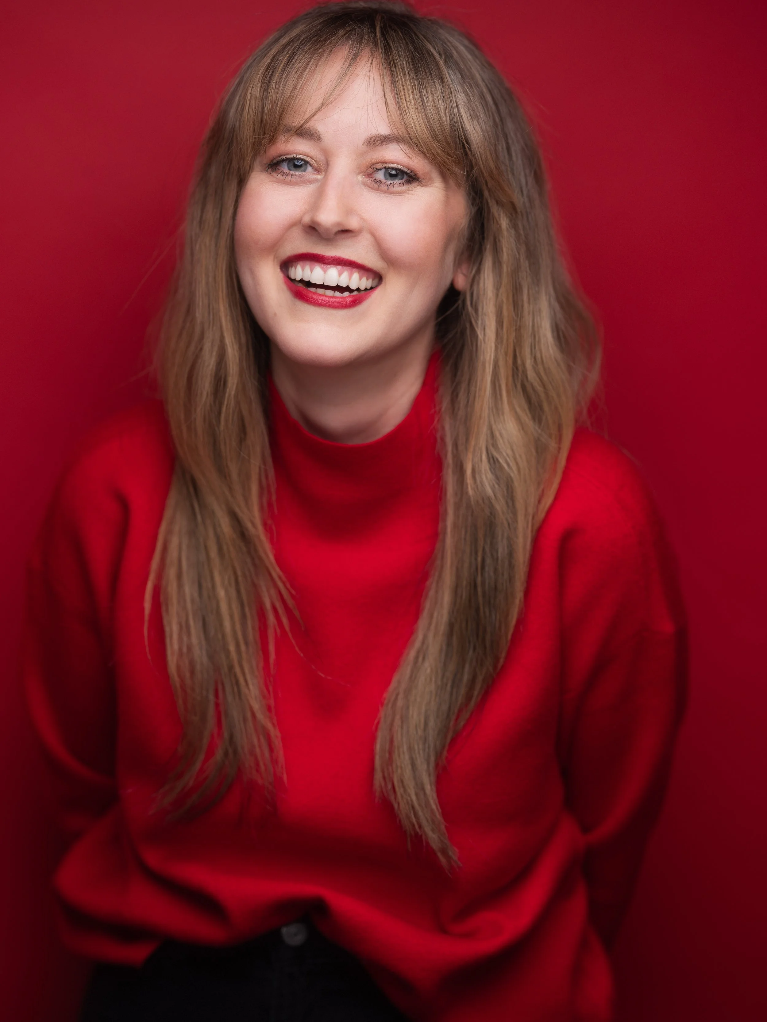 A smiling woman with long blonde hair wearing red lipstick and a red sweater against a red background.