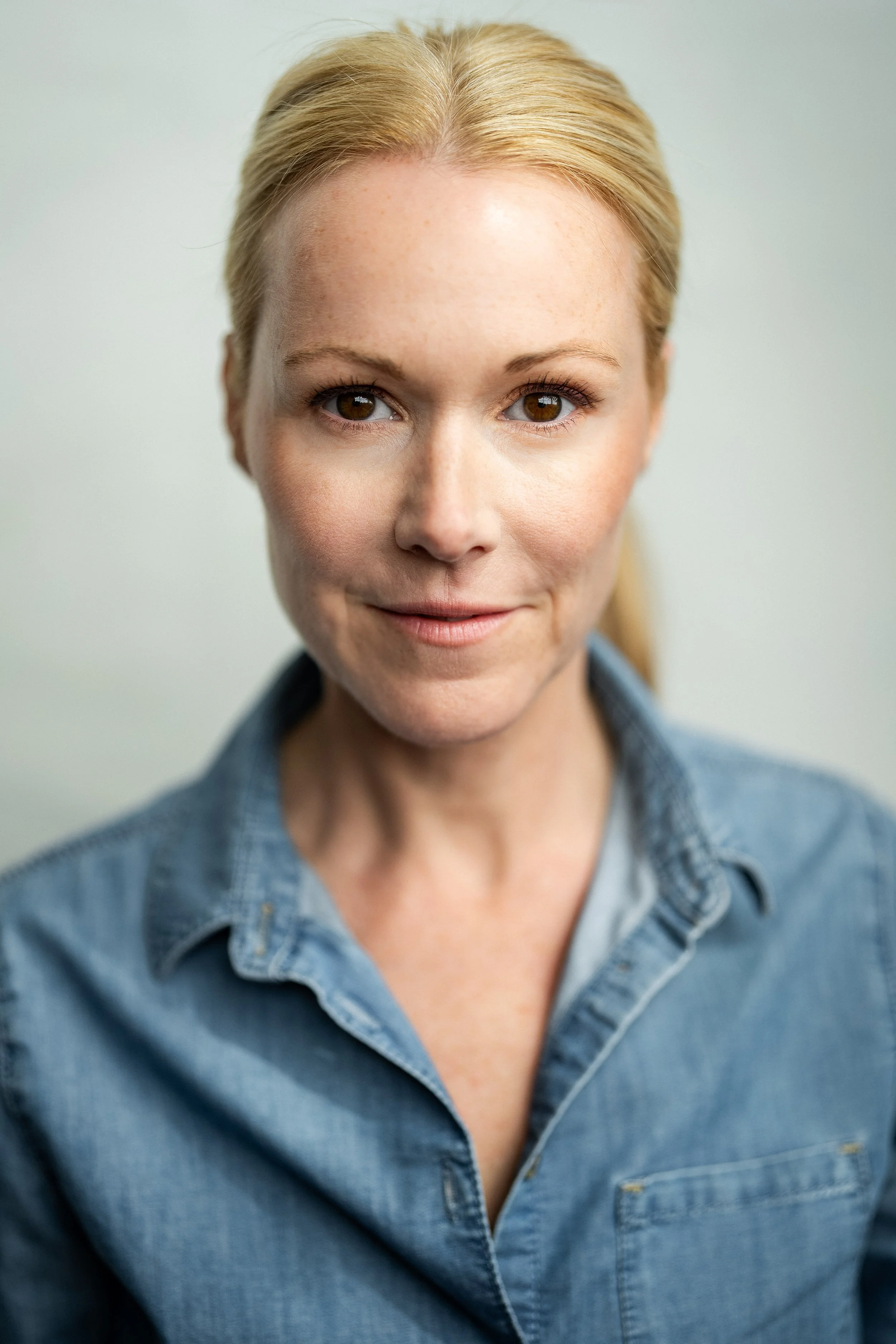 Close-up portrait of a woman with blonde hair, wearing a denim shirt, smiling gently, in front of a neutral background.