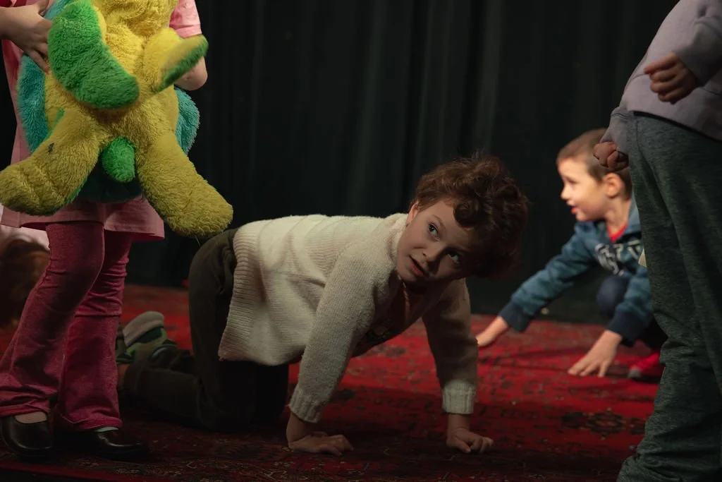 Children participating in a puppet show or performance on a stage with a red carpet, with a dark curtain backdrop.