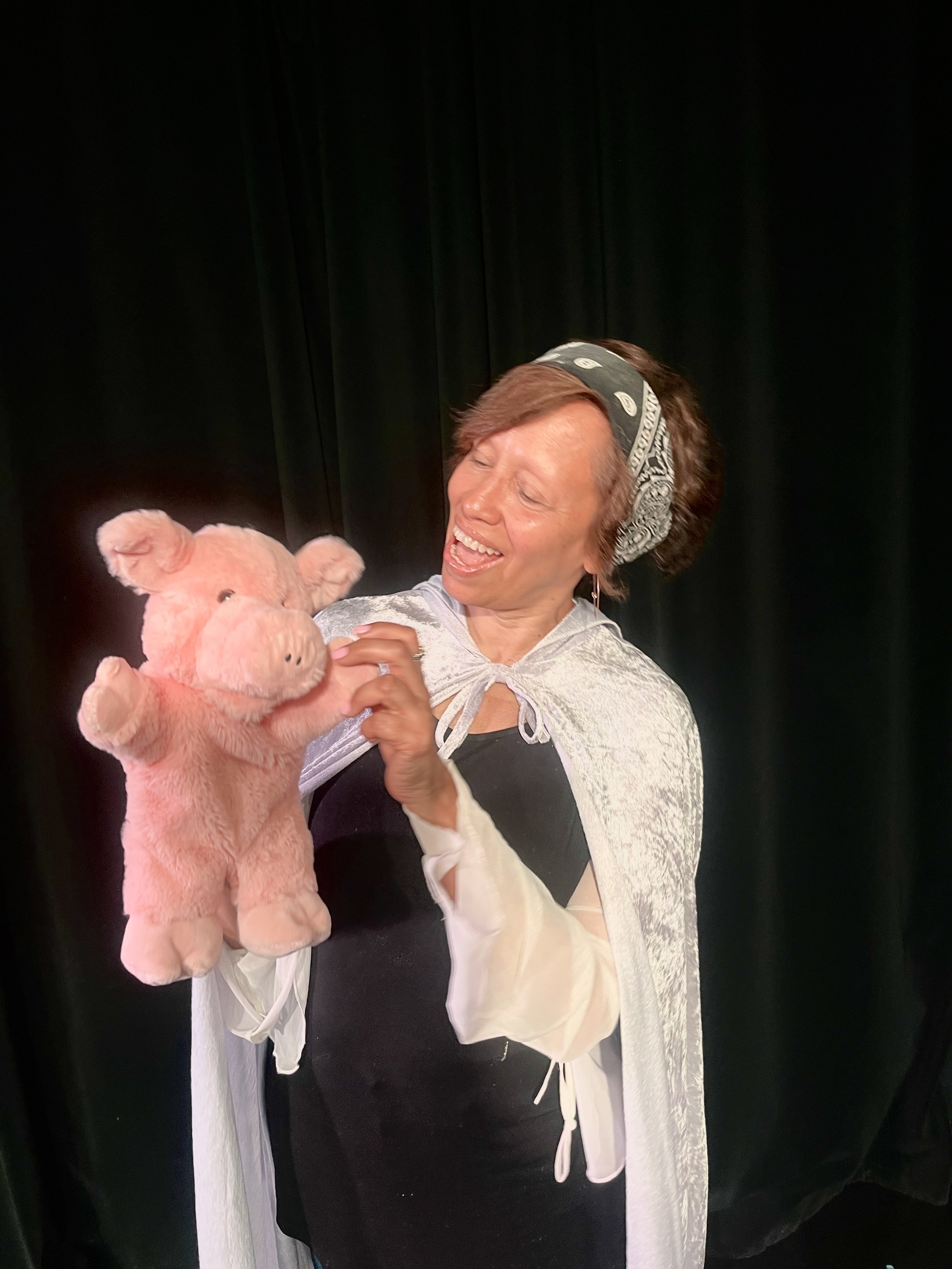 Woman with short brown hair, wearing a bandana and a white shawl, holding a pink stuffed pig, smiling with her eyes closed, standing in front of a black curtain.
