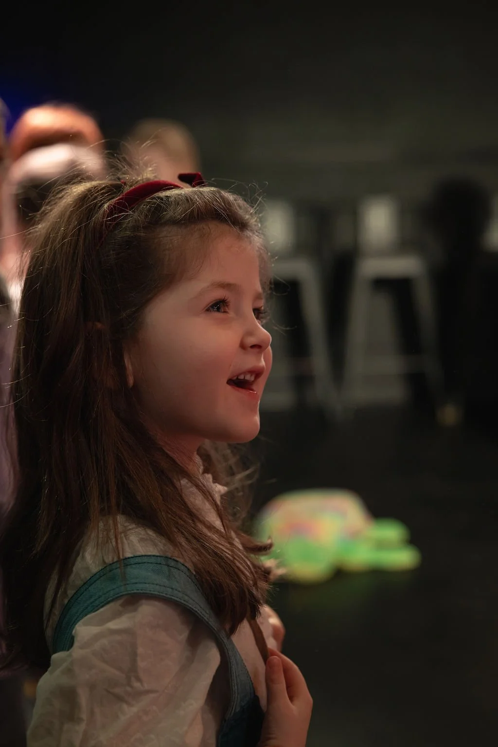 A young girl with brown hair, wearing a beige shirt and a teal backpack, looking forward with a joyful expression in a dimly lit room.