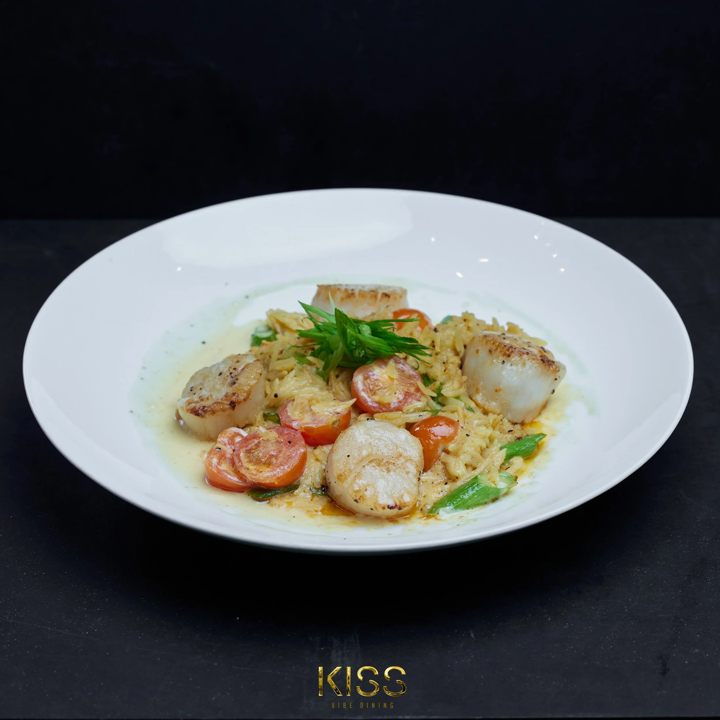 A plate with grilled scallops, cherry tomatoes, green onions, and garnished with green herbs and a creamy sauce.