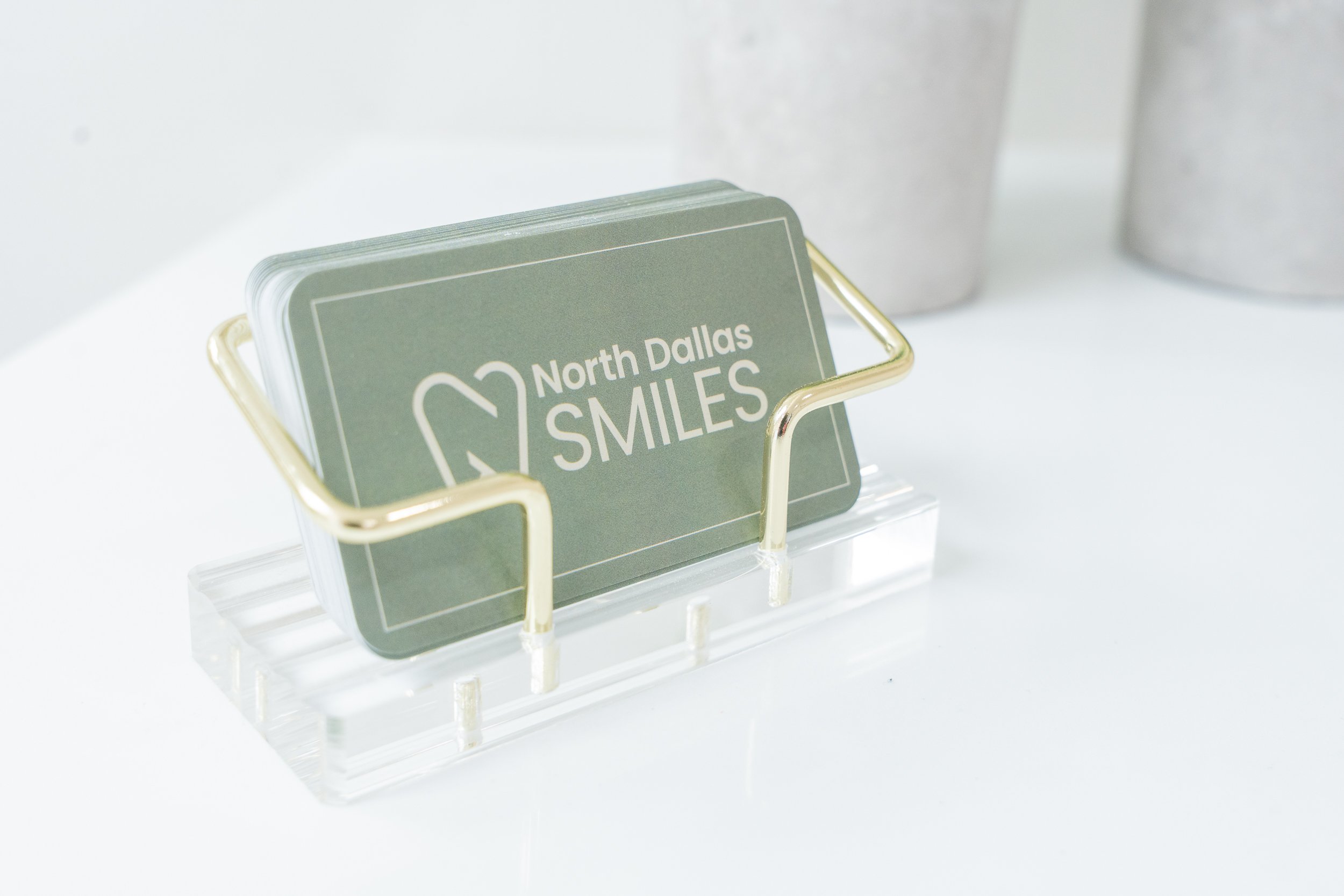 Business cards for North Dallas Smiles in a clear plastic holder on a white surface with blurred containers in the background.