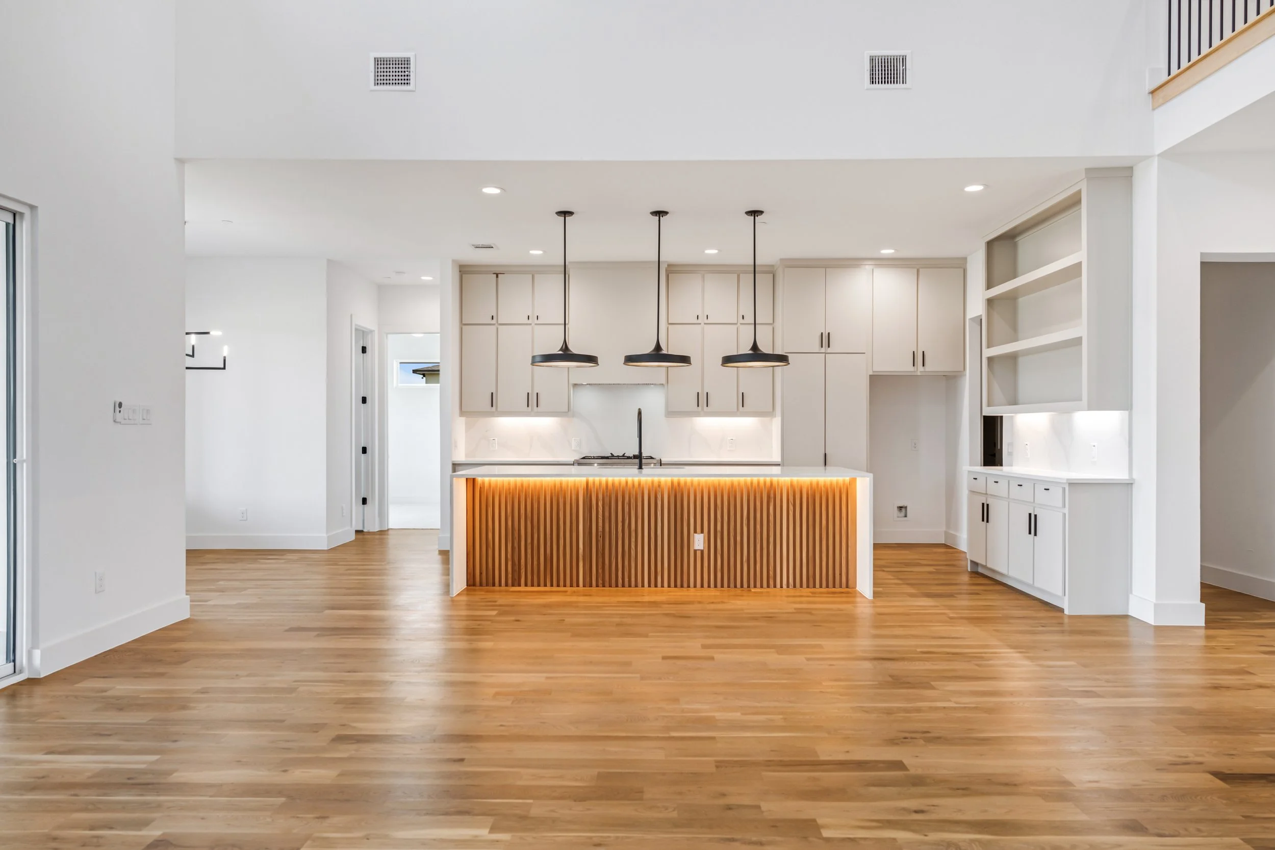 Modern kitchen with white cabinets, wooden island with backlit strip, three black pendant lights, hardwood floors, open shelving, and minimal decor.