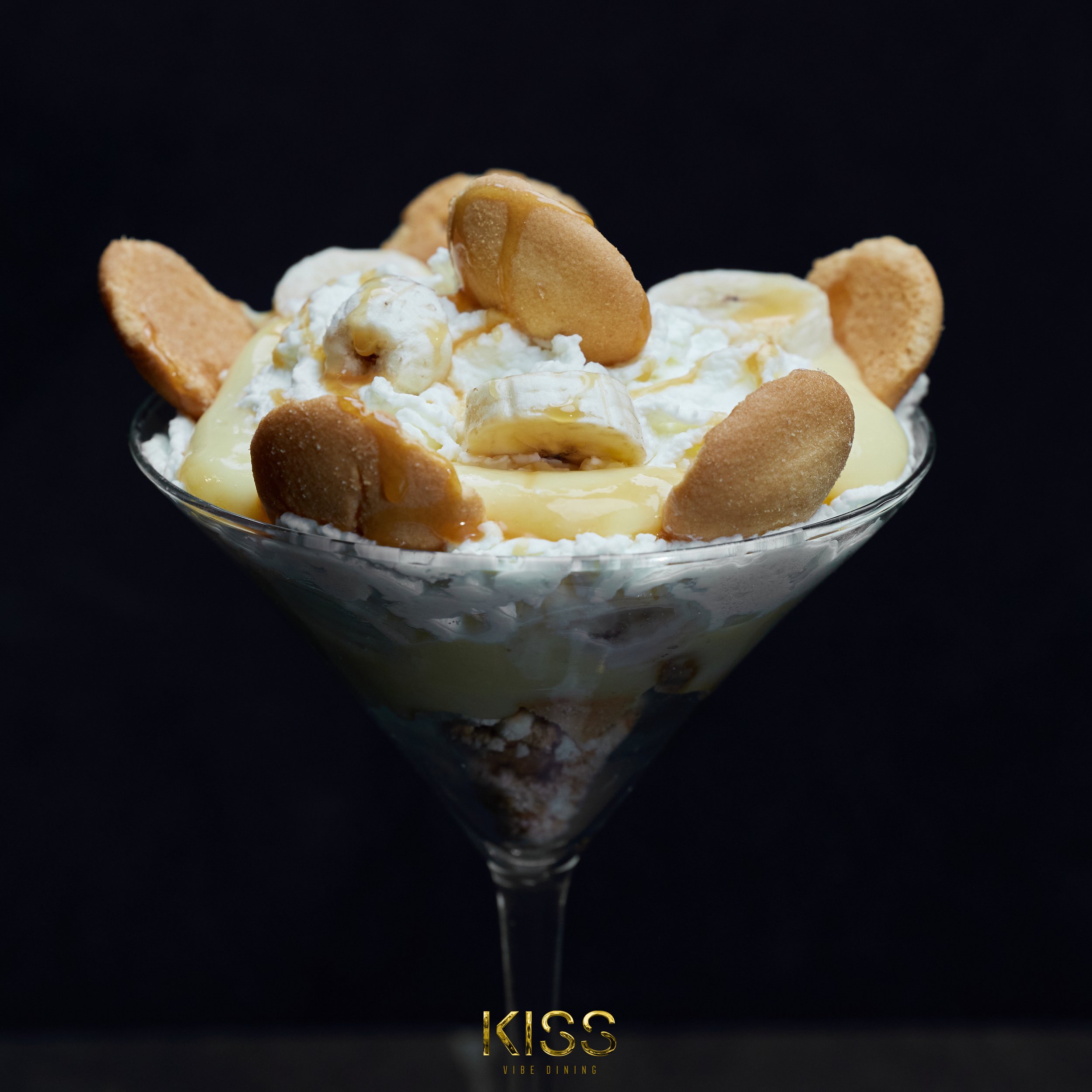 Dessert in a martini glass with whipped cream, banana slices, cookies, and caramel drizzle on top