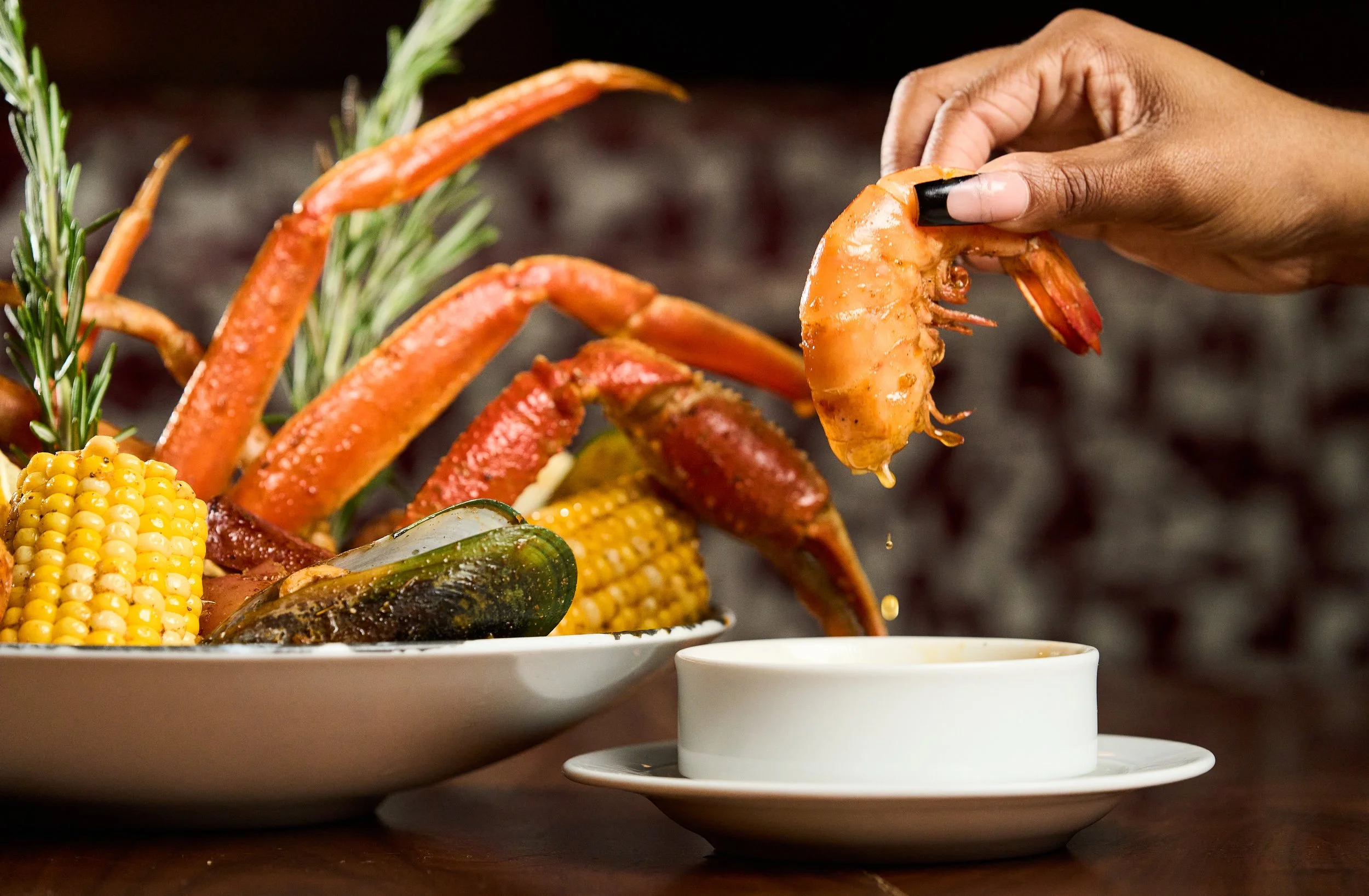 Person holding a cooked shrimp above a white cup on a plate of seafood and corn.