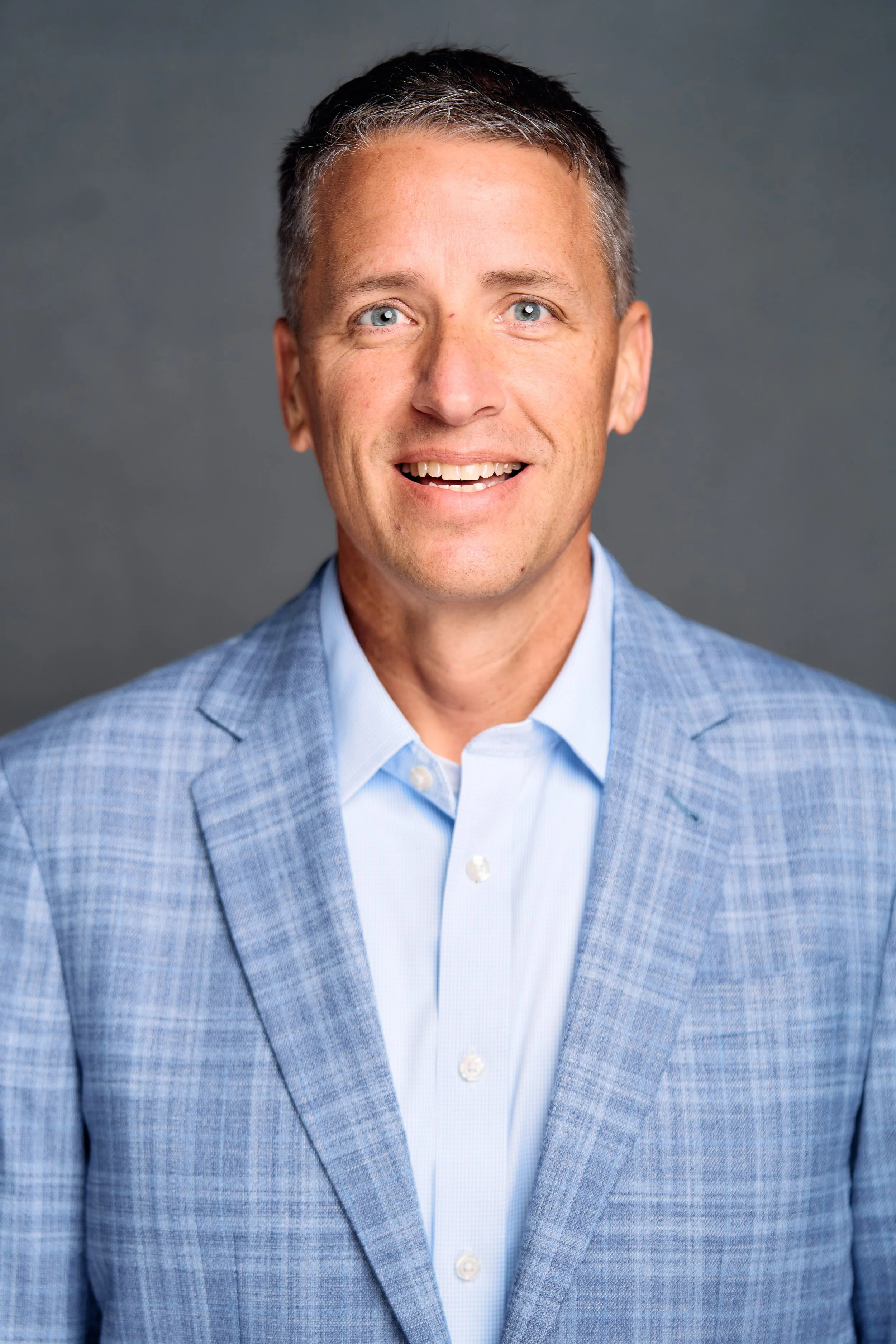 Headshot of a man with short gray hair, blue eyes, smiling, wearing a light blue dress shirt and a blue blazer against a plain gray background.