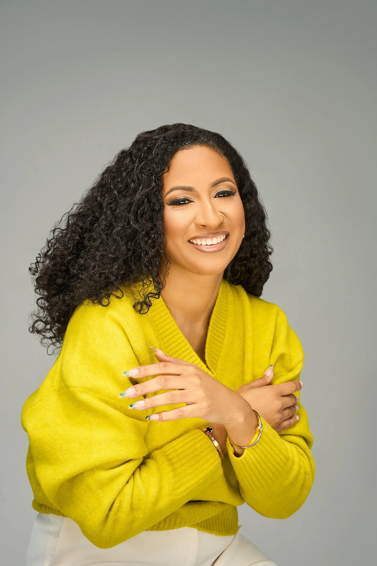 A woman with curly hair, wearing a yellow sweater, smiling and embracing herself against a gray background.