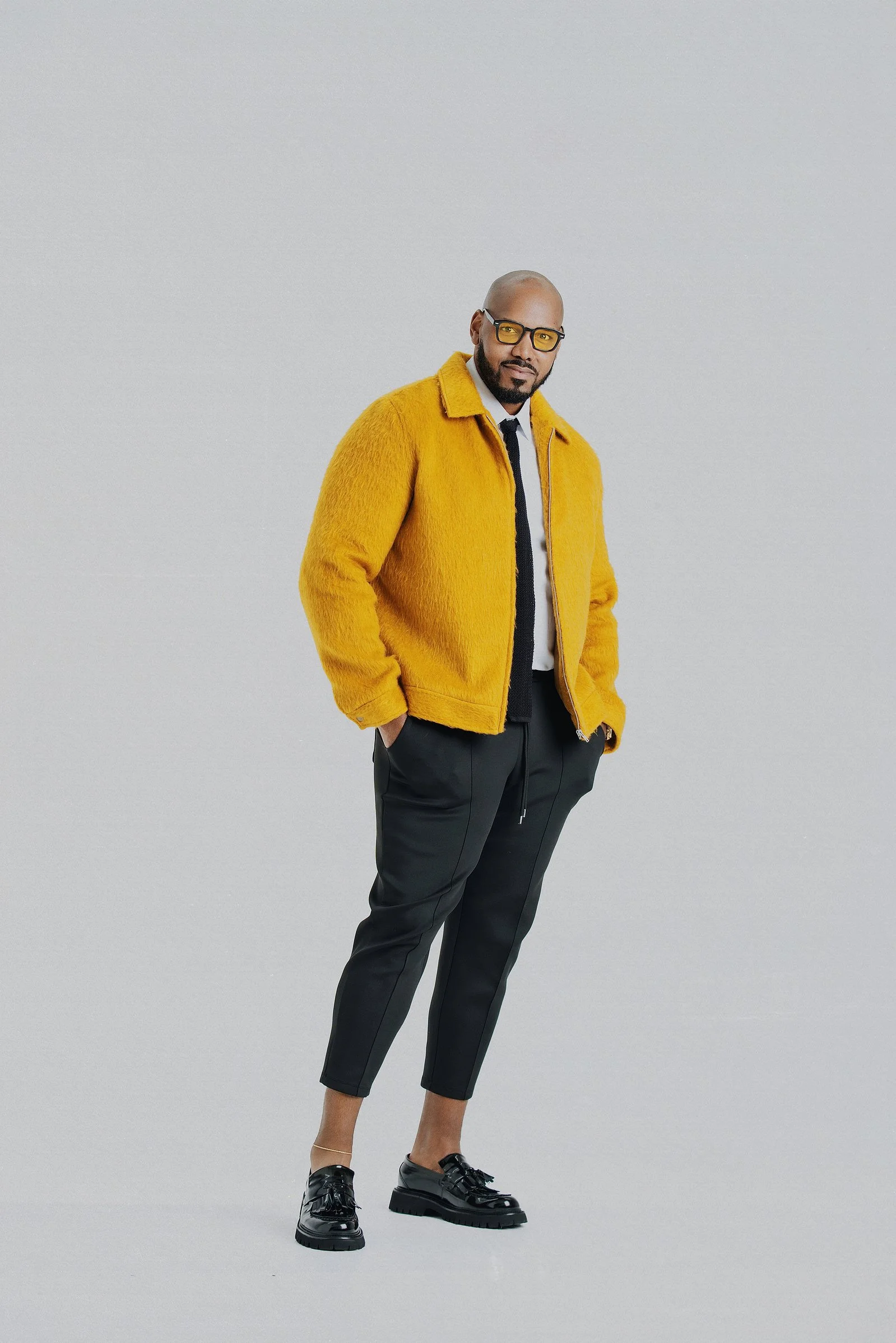 A man with a shaved head, glasses, and a beard stands against a plain light gray background. He is wearing a bright yellow fuzzy jacket, black pants, and black shoes, and is looking at the camera.