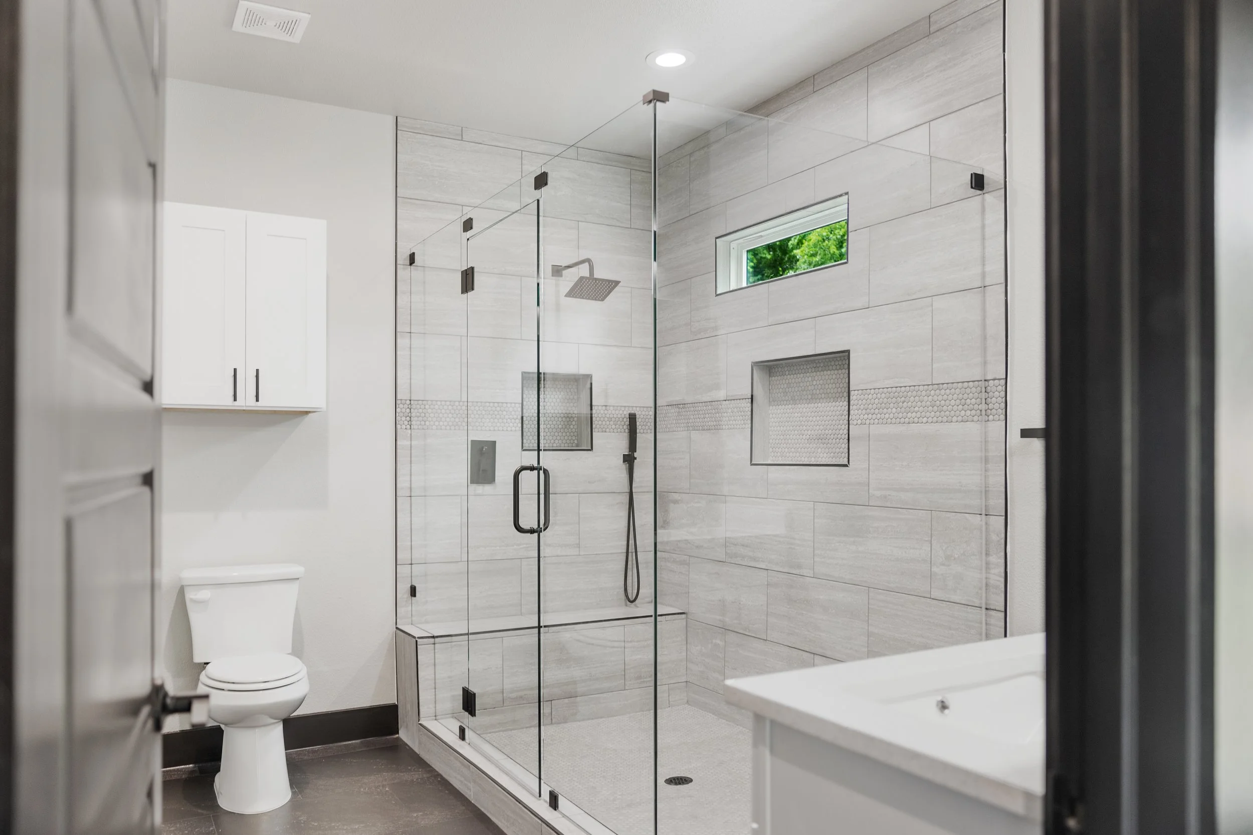 Modern bathroom with a glass-enclosed shower, small window, white toilet, and white cabinet.