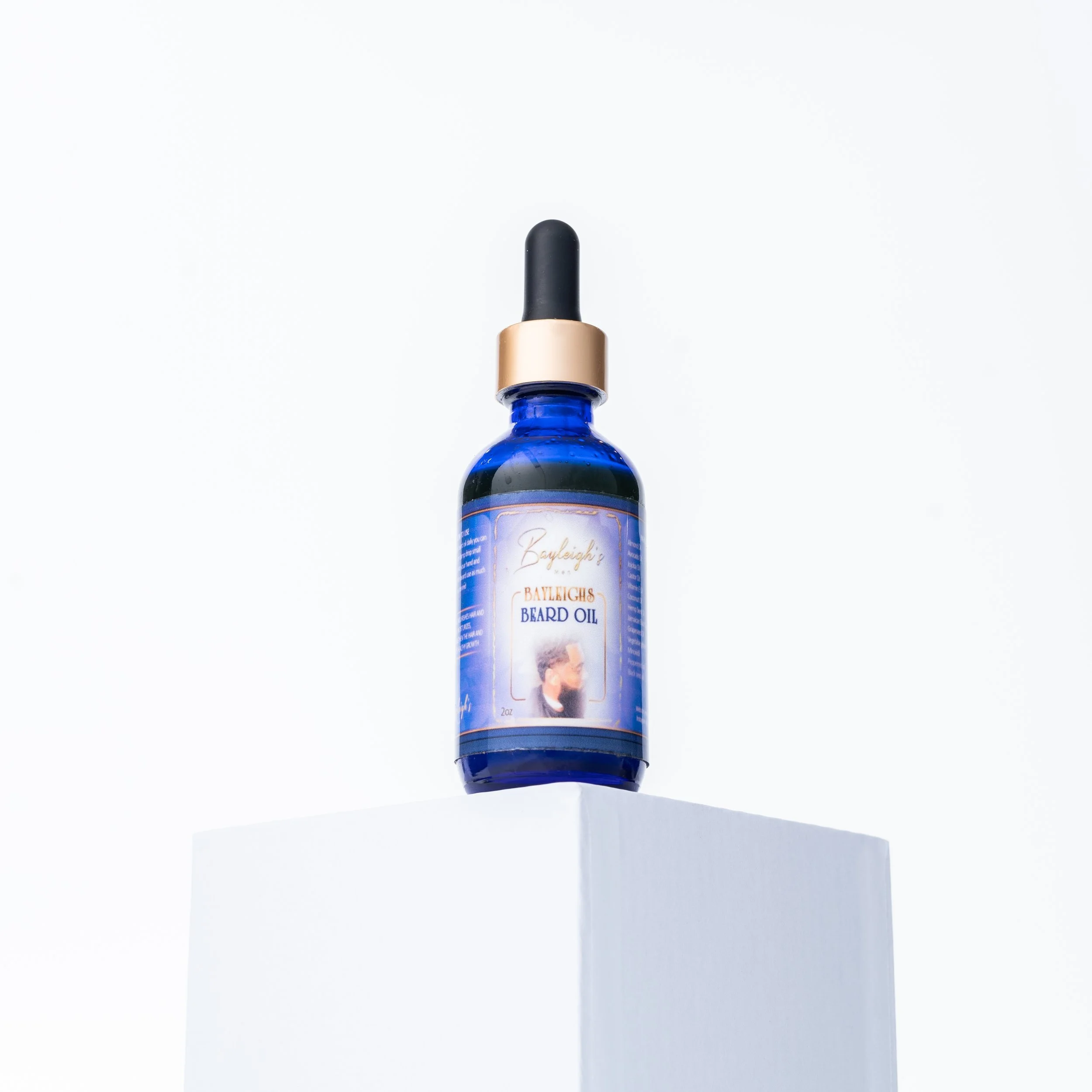 A blue bottle of beard oil with a black dropper cap, placed on a white pedestal against a plain white background.