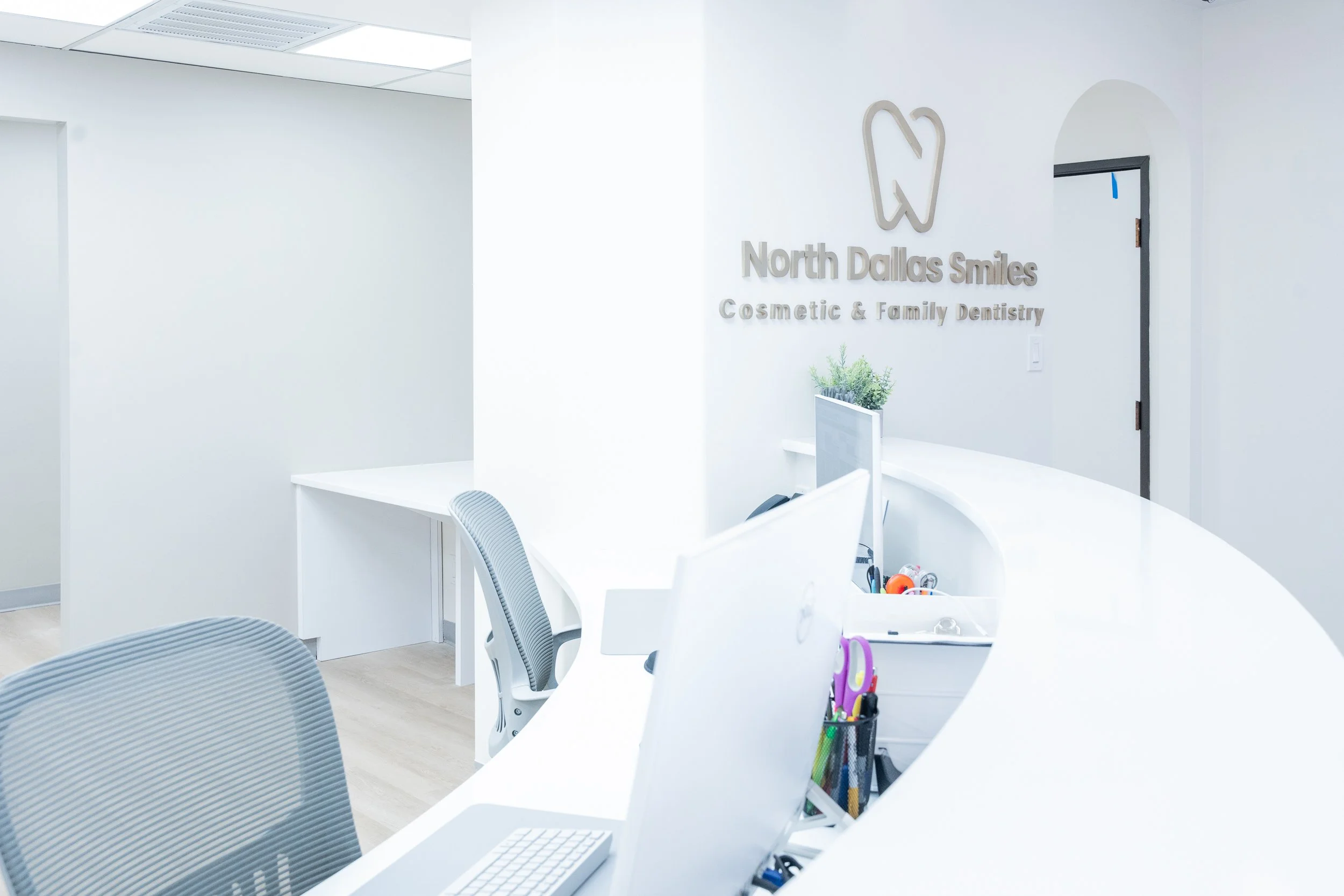 Reception area of North Dallas Smiles, a cosmetic and family dentistry office, with a white curved counter, computer, office supplies, and a sign on the wall displaying the practice's name and logo.