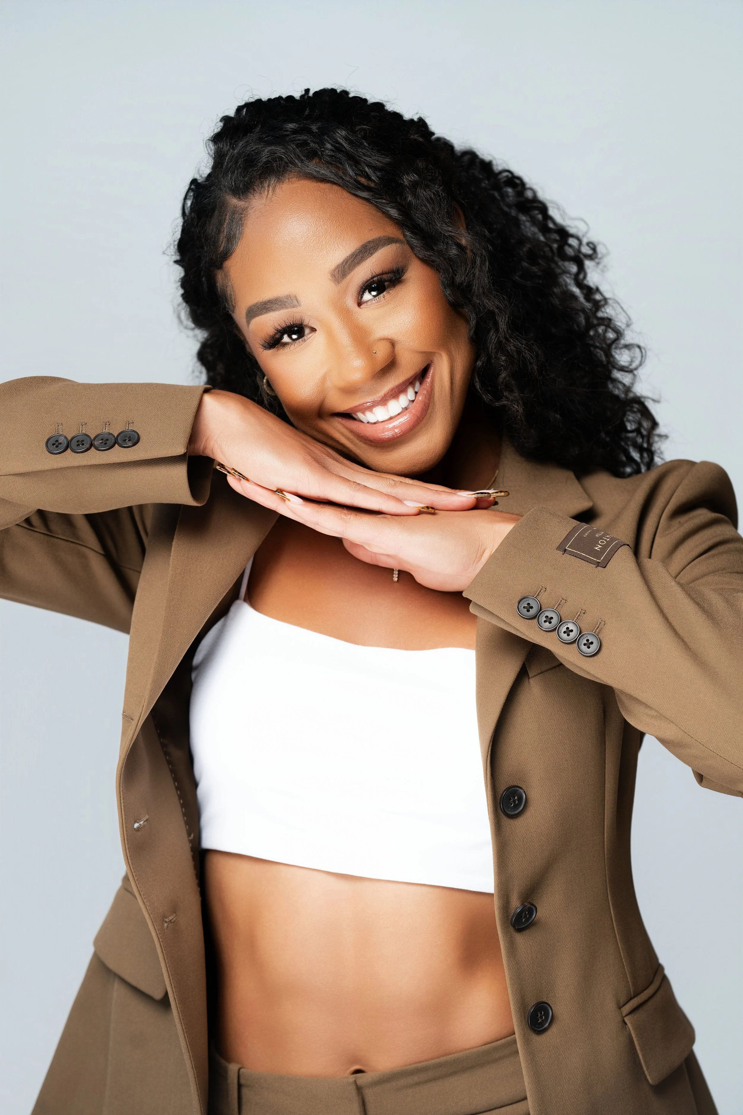 A smiling woman with curly black hair, wearing a tan blazer and white crop top, posing with hands under her chin against a plain light gray background.
