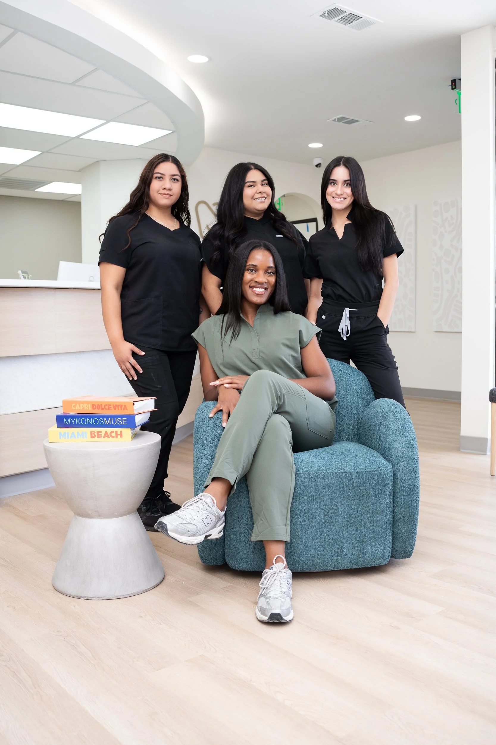 Four women in medical scrubs pose in a modern clinic lobby, with a chair, colorful books, and light wood flooring.