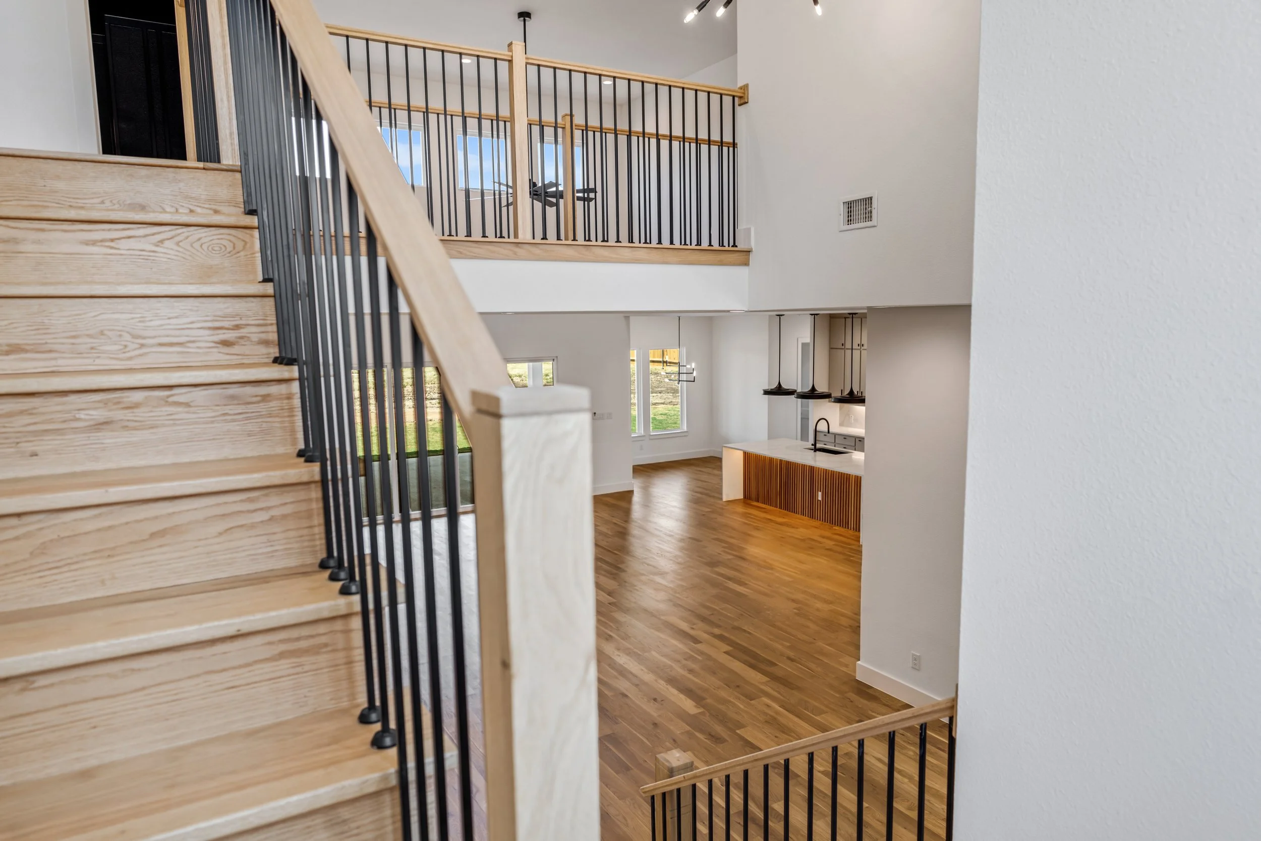 Interior view of a modern multi-level home with wooden stairs and black metal railing, open-concept kitchen with pendant lights, and large windows letting in natural light.