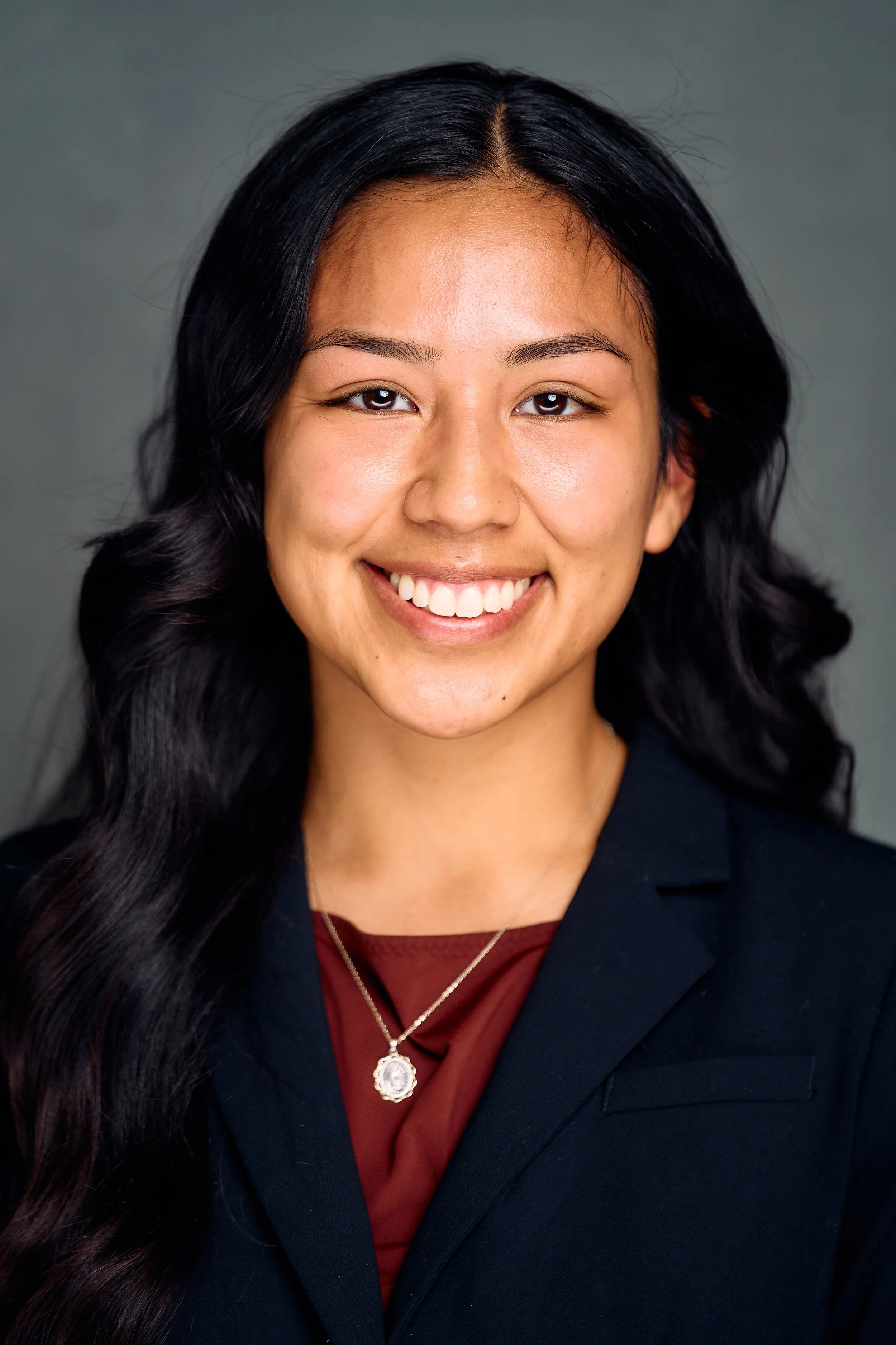 A professional woman with long black hair, smiling, wearing a black blazer, maroon top, and a pendant necklace, against a gray background.