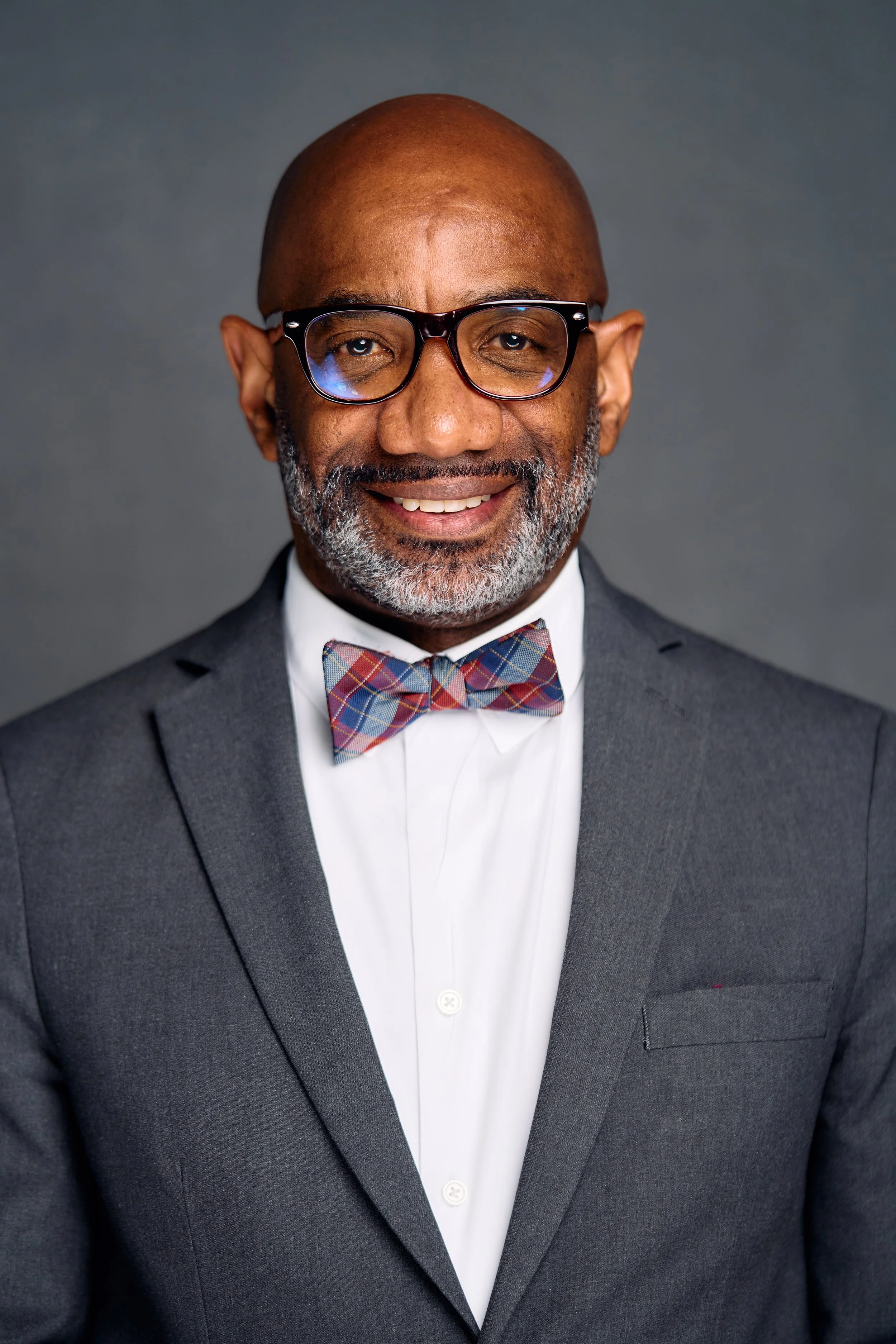 A professional portrait of a middle-aged African American man wearing glasses, a suit, a white shirt, and a plaid bow tie, smiling against a gray background.