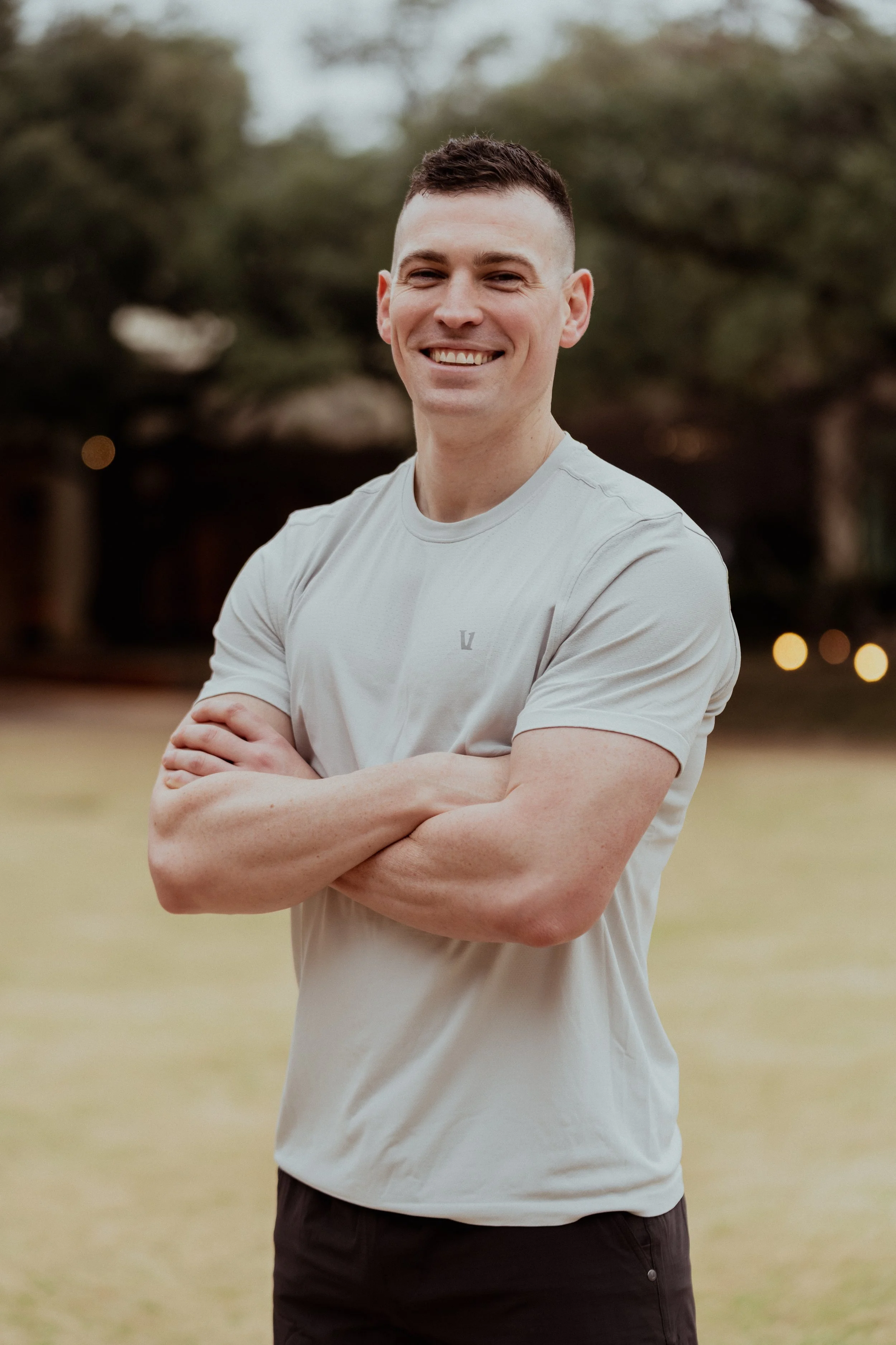 A smiling young man with short dark hair and a fit build, standing outdoors with arms crossed, wearing a light-colored athletic shirt and dark shorts, in a park or grassy area with trees in the background.