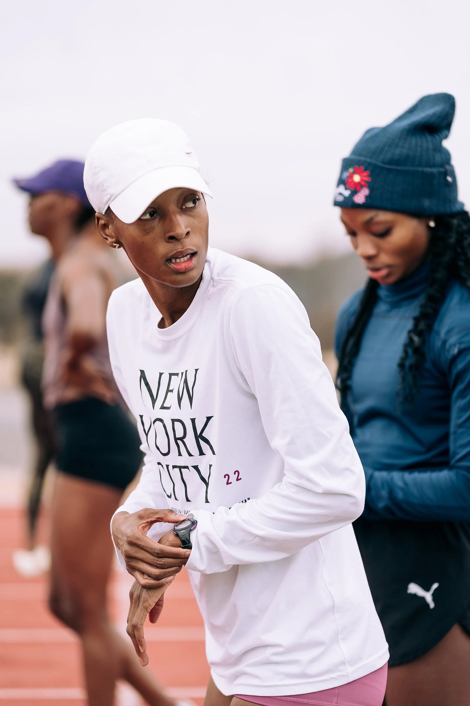 A woman with a white baseball cap and white shirt that says 'New York City' is looking at her watch while preparing for a race on a track. Other women are in the background.