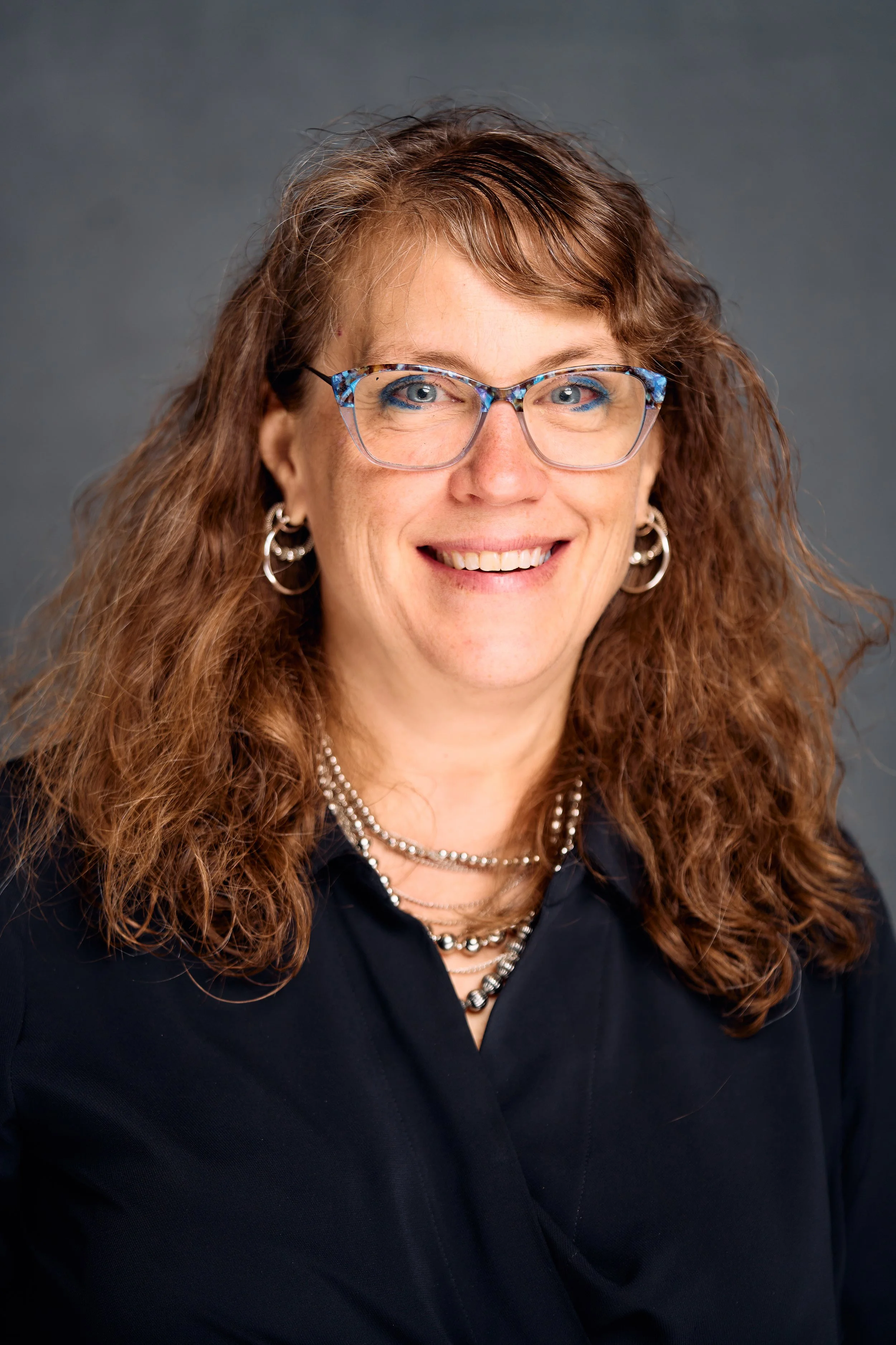 A woman with glasses, curly red hair, and silver jewelry smiling against a dark background.