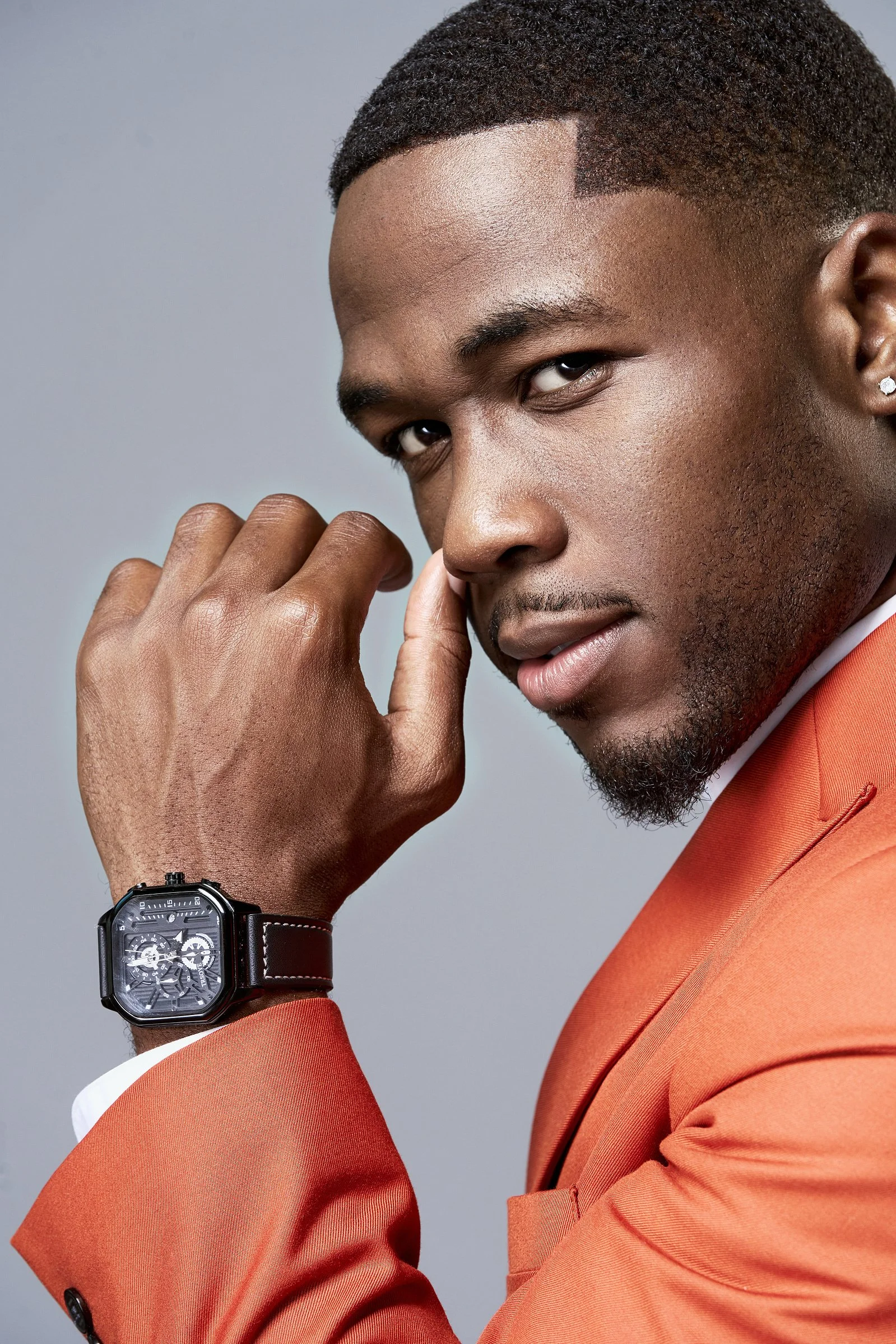 A young man with dark skin and short hair, wearing an orange suit, poses with his hand near his face, showing a black wristwatch with a complex dial.