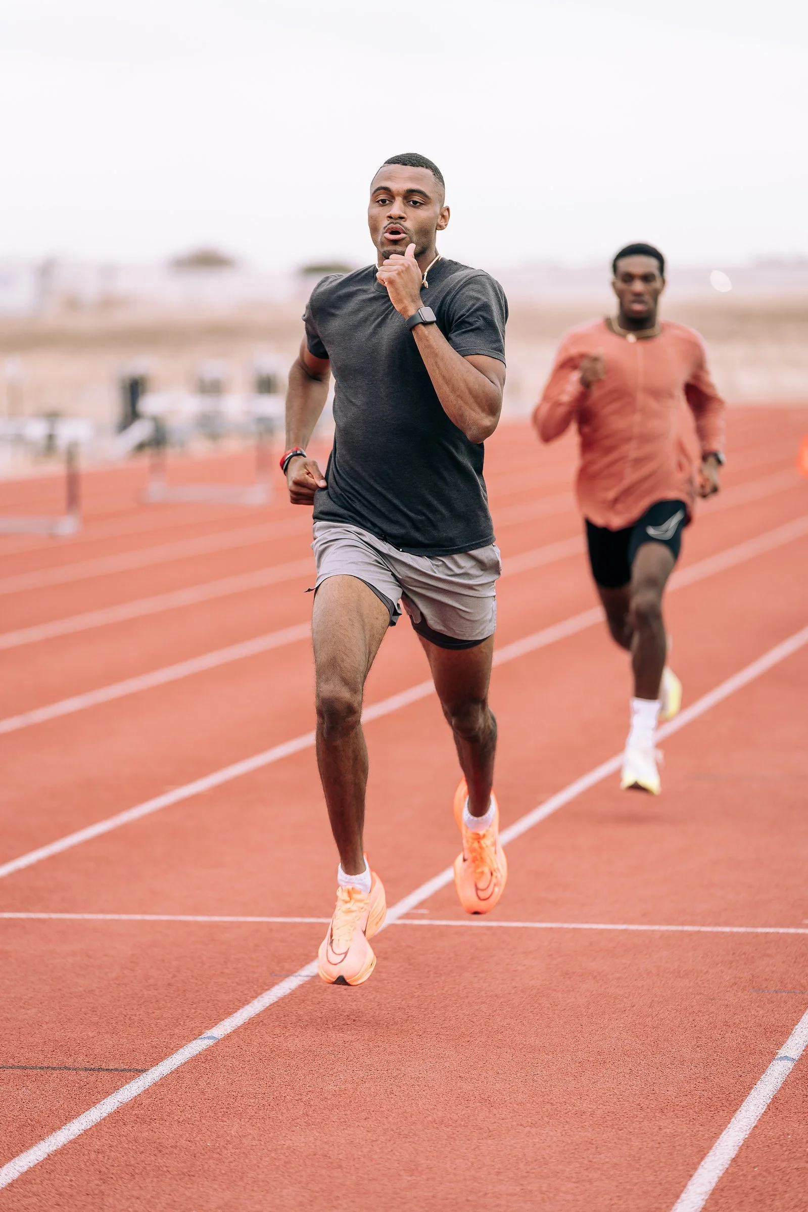 Two men running on a red outdoor track, focused on the man in front who is wearing a black T-shirt, gray shorts, and orange running shoes, with another man in a burnt orange shirt and black shorts running behind him.