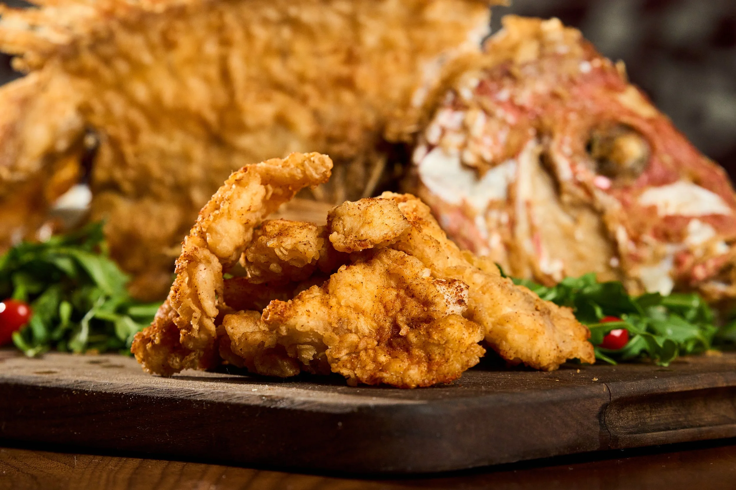 Fried chicken pieces on a wooden serving board, with a partially visible cooked fish and some greens with red berries in the background.