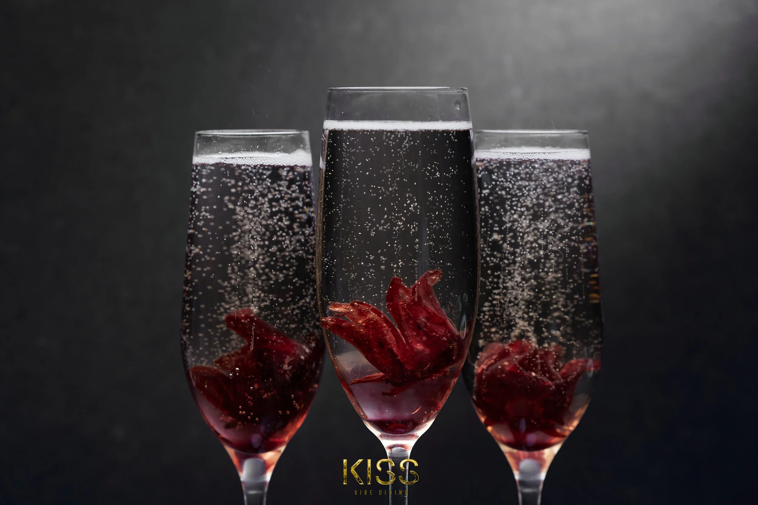 Three champagne flutes filled with dark red liquid, each containing a whole cooked lobster at the bottom, against a dark background with bubbles, with the word 'KISS' in gold lettering at the bottom.