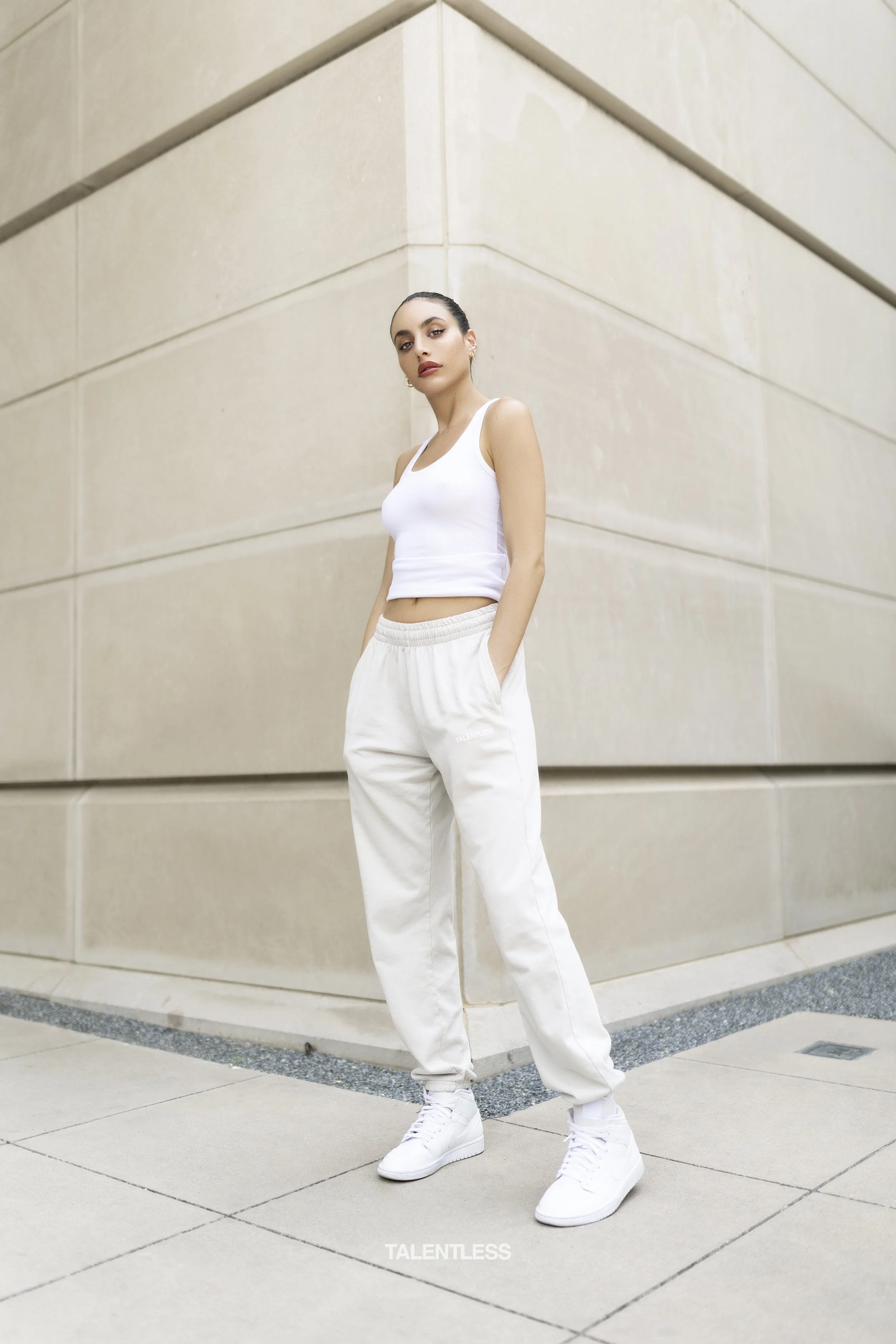 A young woman with dark hair tied back, wearing a white tank top, white sweatpants, and white sneakers, standing outdoors against a beige stone building wall with her hands in her pockets.