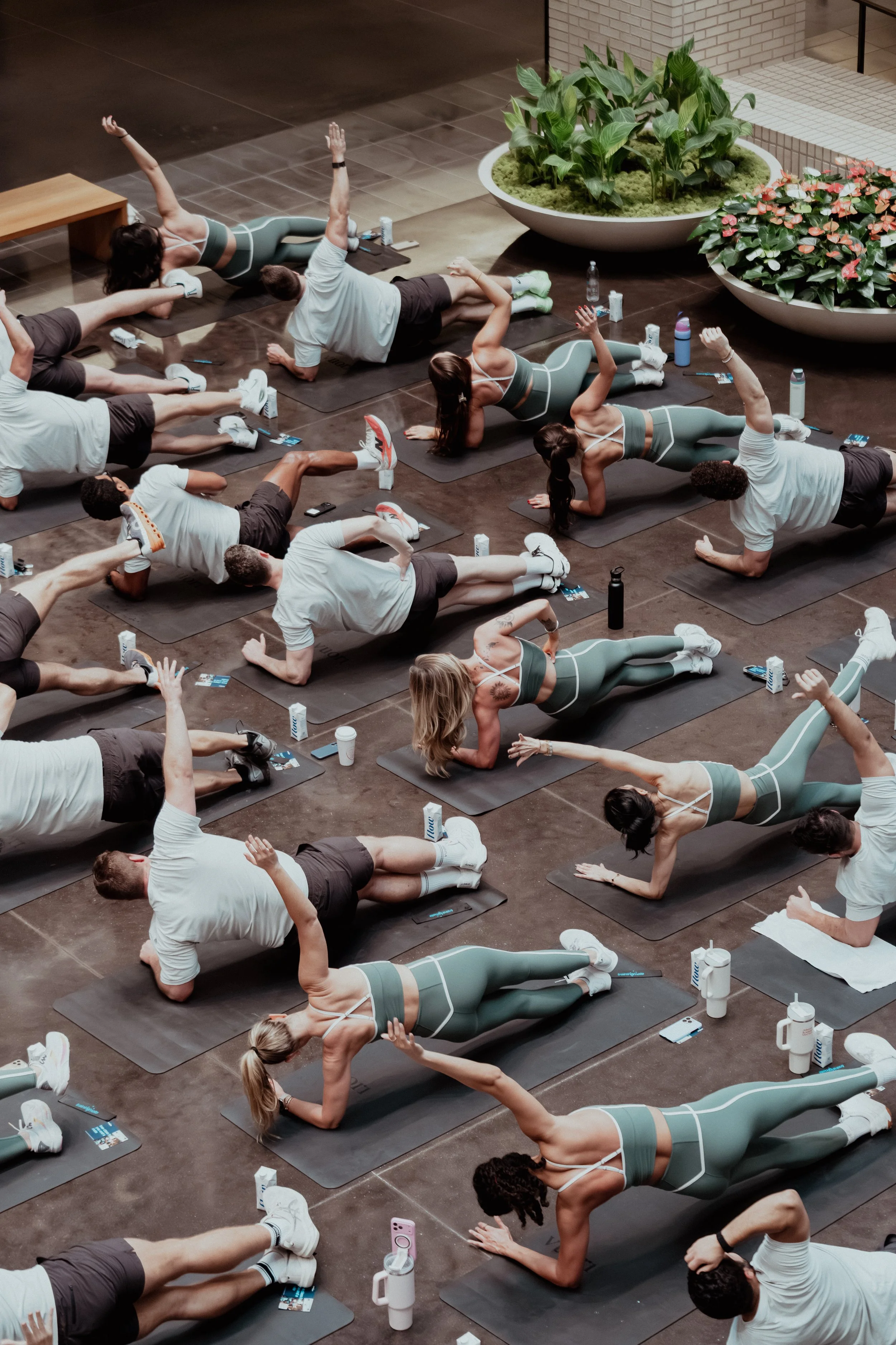 Group of people participating in a fitness class on yoga mats, performing side planks, in an indoor setting with plants and water bottles on the floor.