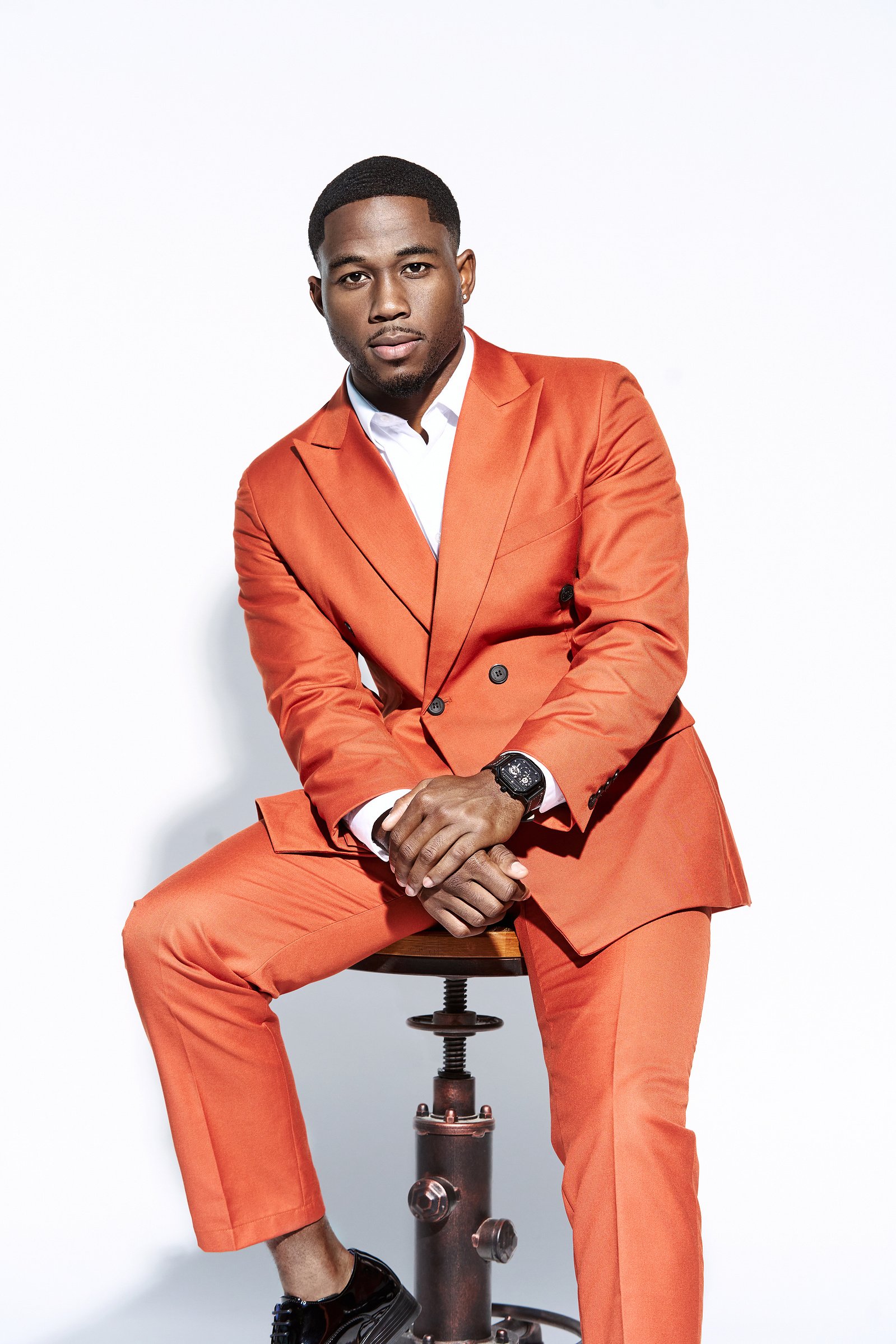 A young man sitting on a wooden stool against a white background, wearing an orange suit, white shirt, black watch, and black shoes.