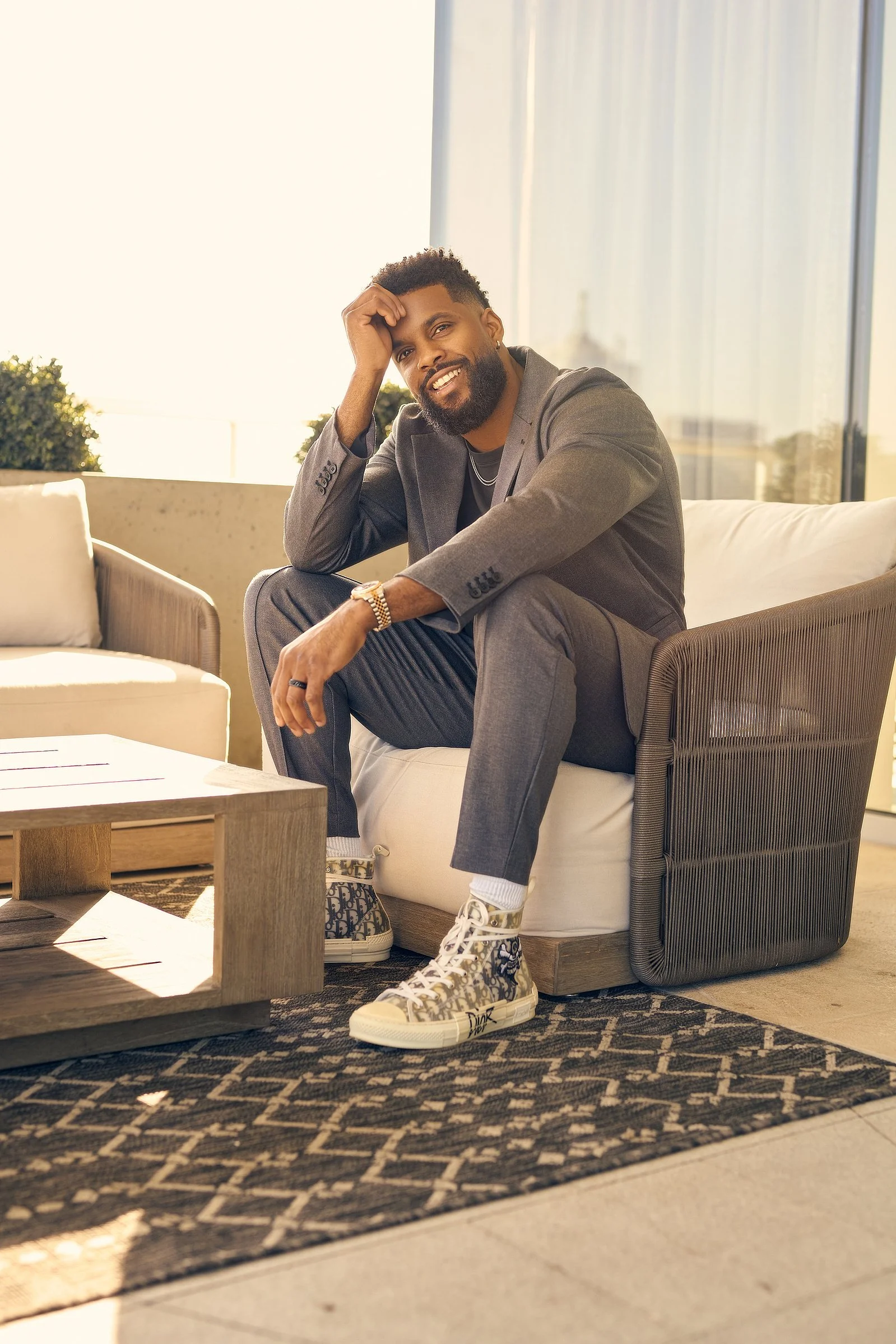 A man in a gray suit sitting on a modern outdoor patio sofa, smiling, with sneakers, in sunlight, in front of a glass window.