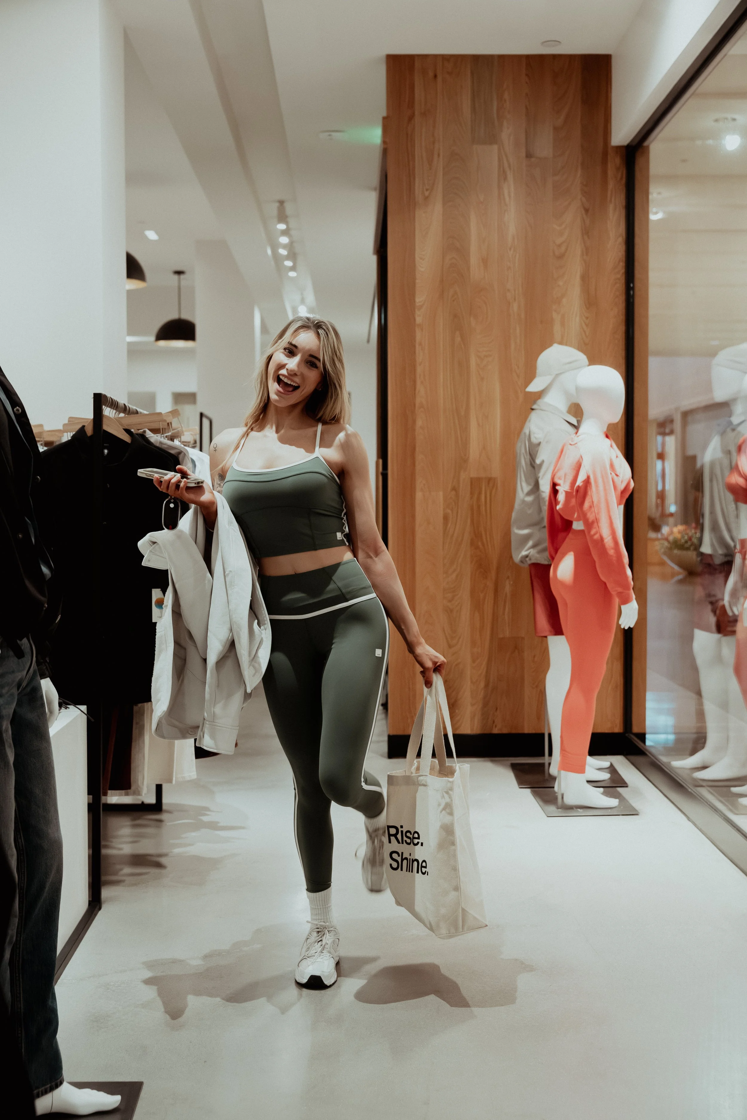 A woman with blonde hair wearing a green athletic outfit, holding a shopping bag that says 'Rise. Shine.' in a store aisle.