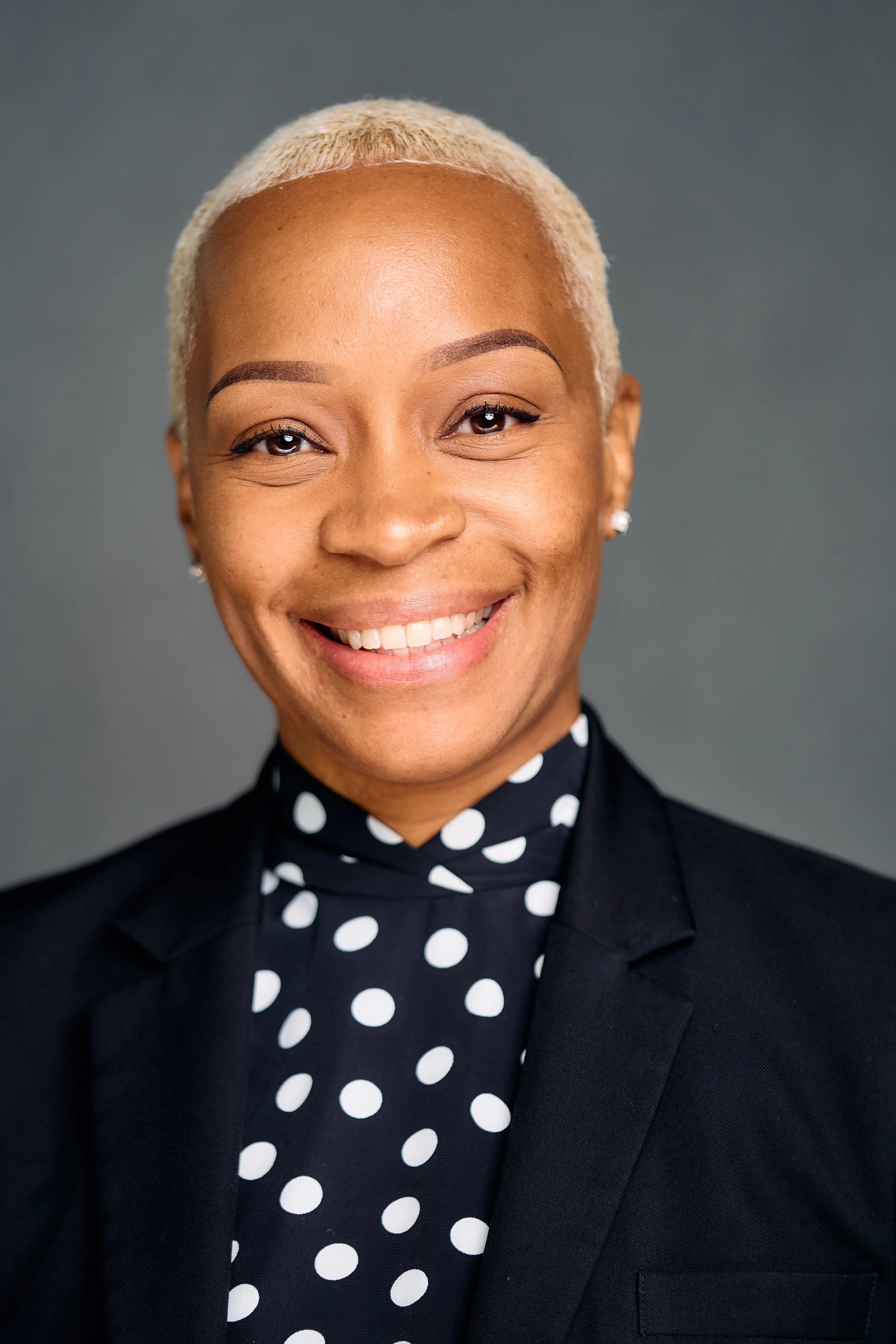 A woman with short, bleached blonde hair, wearing a black blazer and polka dot blouse, smiling against a gray background.
