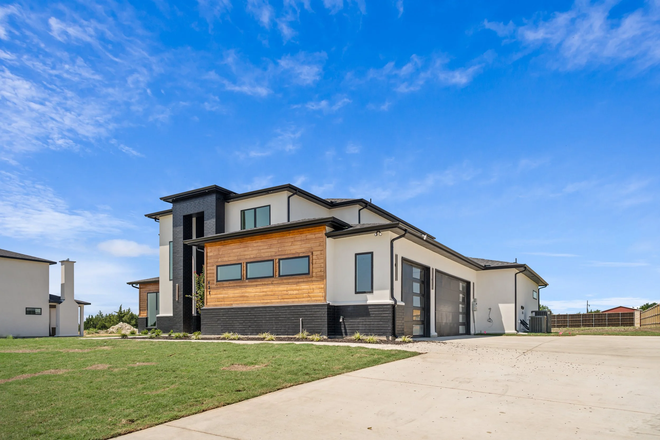 Modern two-story house with a combination of black, white, and wood exterior, large windows, and a spacious concrete driveway under a blue sky with scattered clouds.