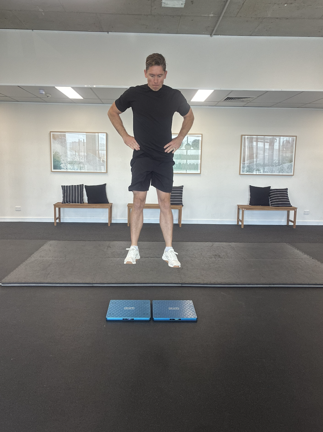 A man in black workout clothing standing on a gym floor, looking down at two blue digital scales  (force plates) placed on the ground in front of him.