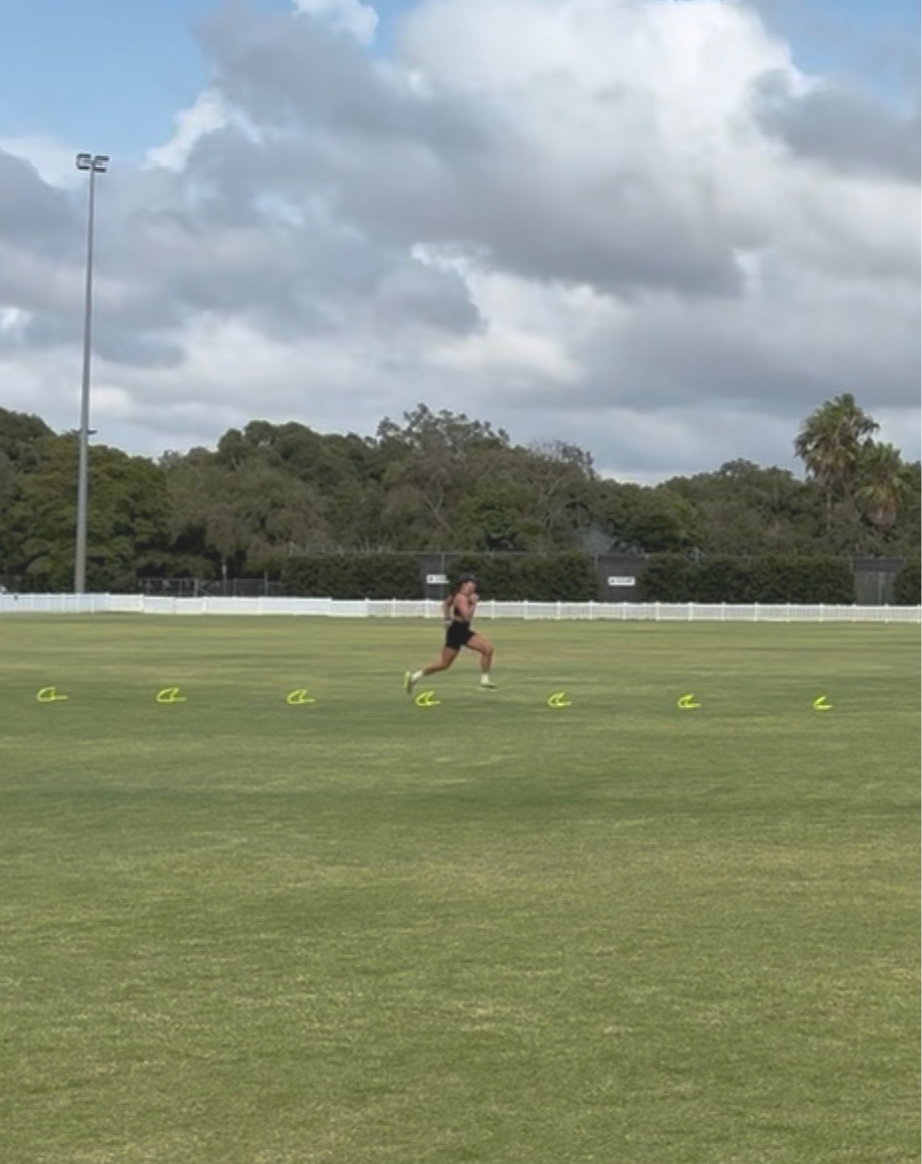 A person is running in an athletic training field marked with several small yellow hurdles, with trees and cloudy sky in the background.