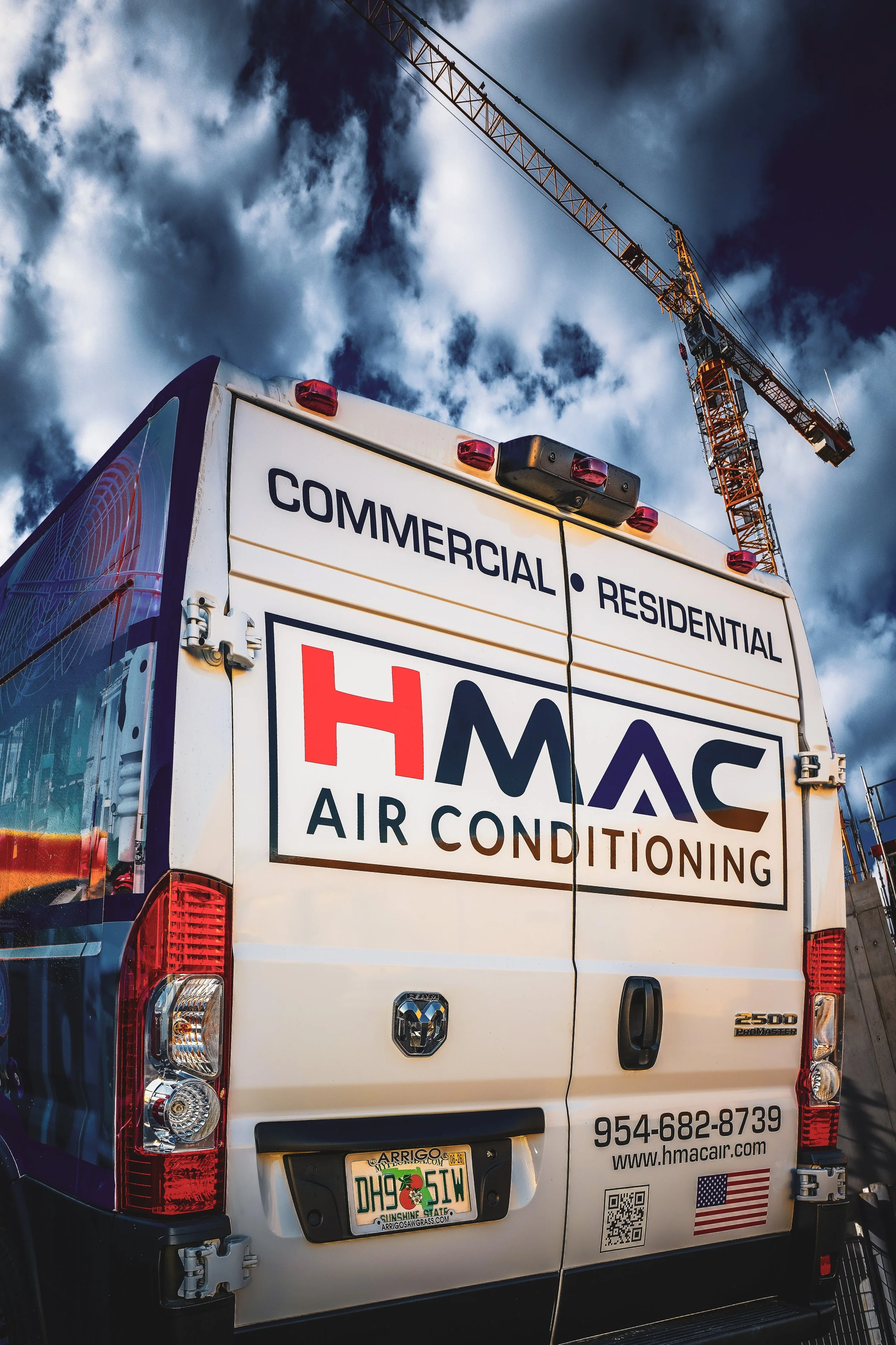 The back of a white commercial van advertising air conditioning services, with a tall construction crane and a cloudy blue sky in the background.