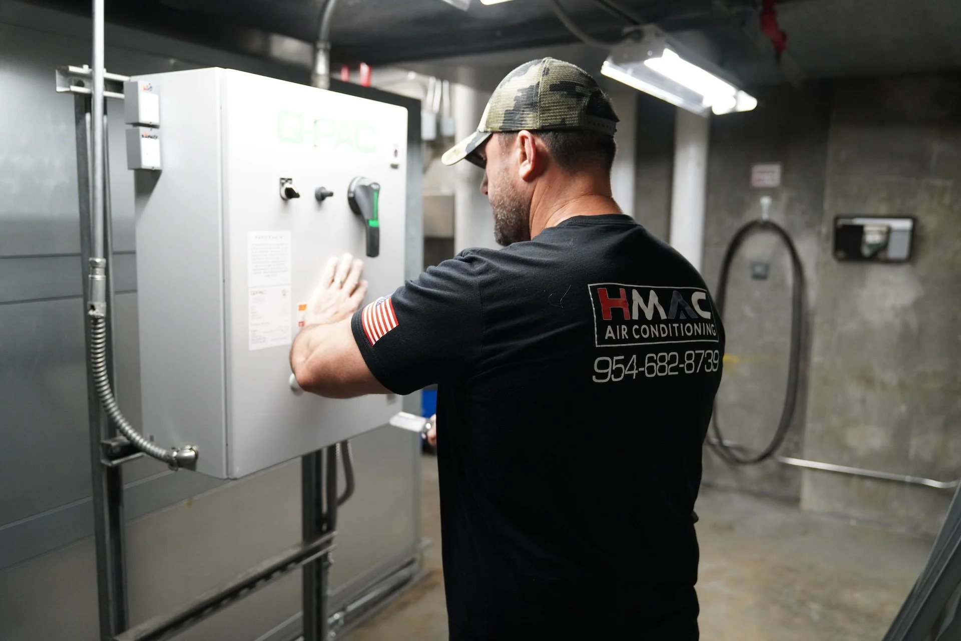 A man wearing a black t-shirt with 'HMAC Air Conditioning' logo and a camouflage cap, working on an electrical panel in an industrial setting.