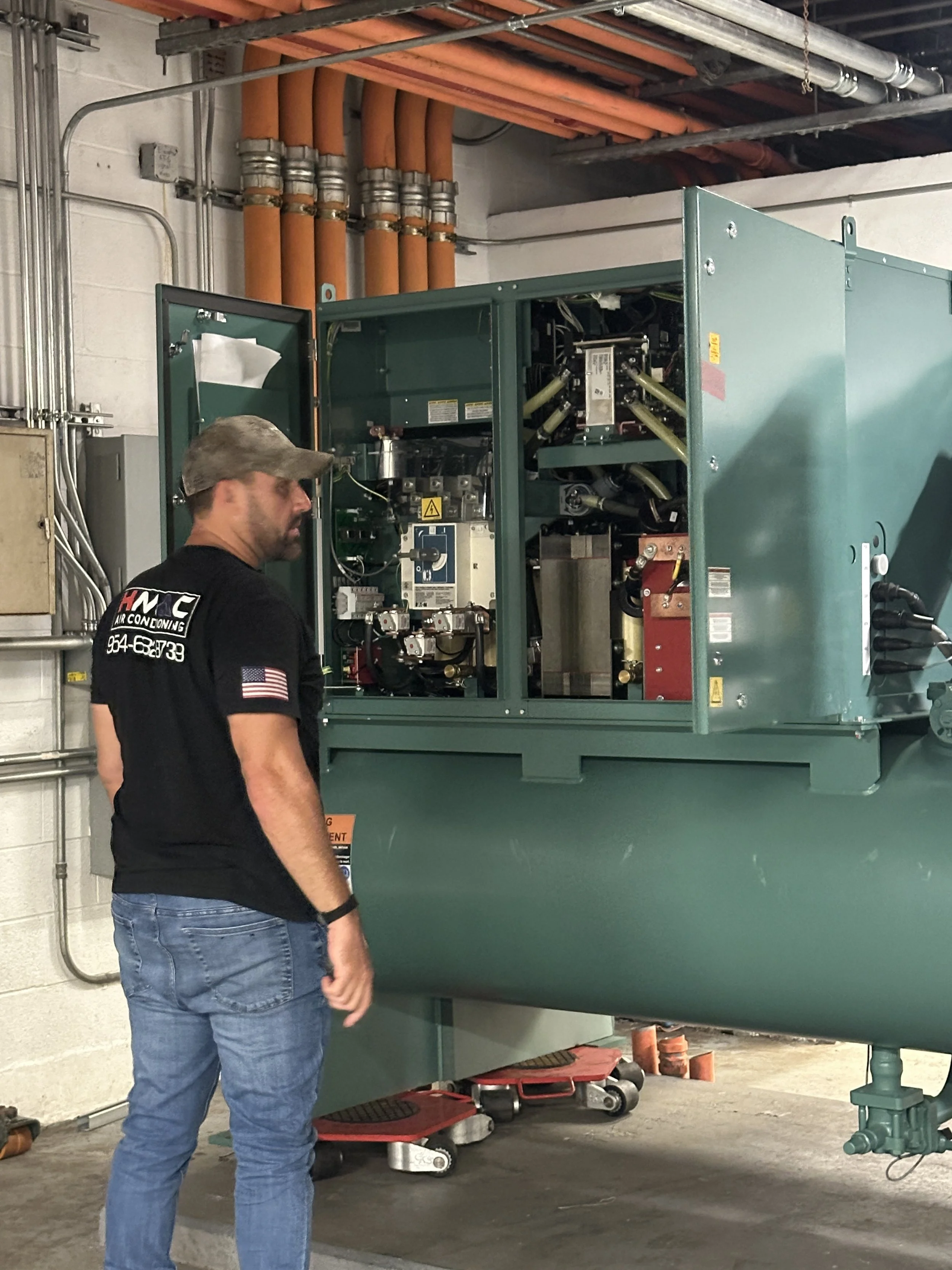 A man wearing a black T-shirt and jeans, with a camouflage cap, standing next to a large green industrial machine in a room with pipes and electrical wiring.