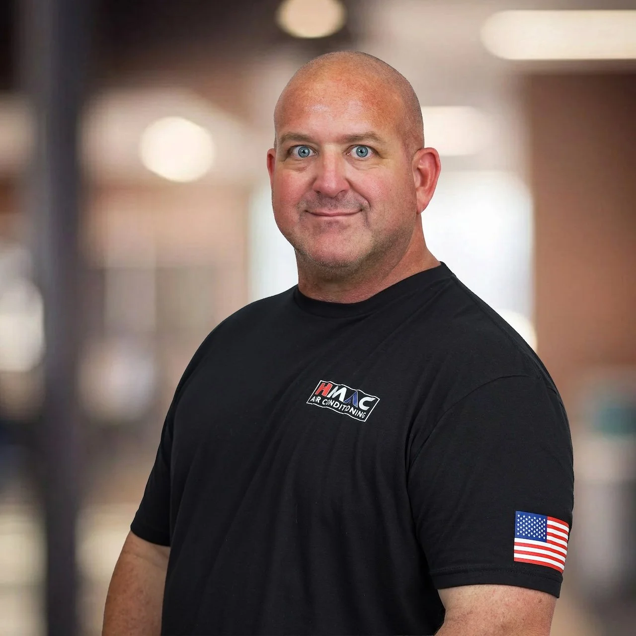 A bald man with blue eyes and facial hair smiling, wearing a black shirt with a logo that reads 'ACME Air Conditioning,' standing in front of a blurred indoor background.