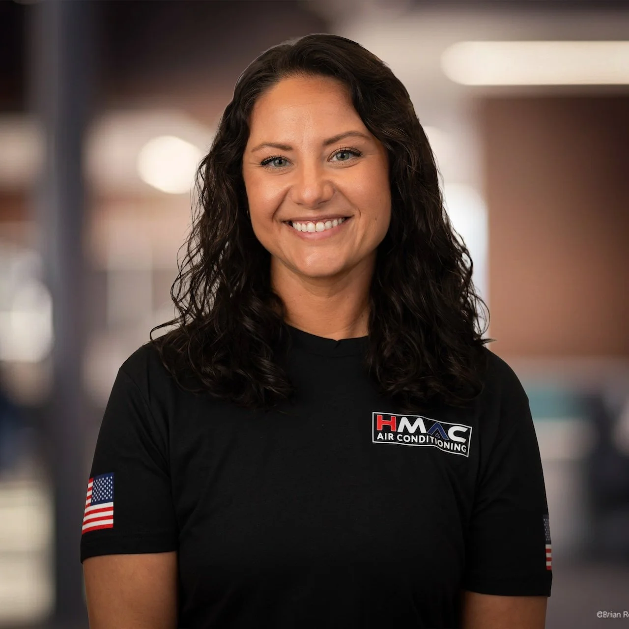 A smiling woman with dark curly hair wearing a black t-shirt with 'HMAC Air Conditioning' logo and American flag patches on sleeves, standing indoors with blurred background.