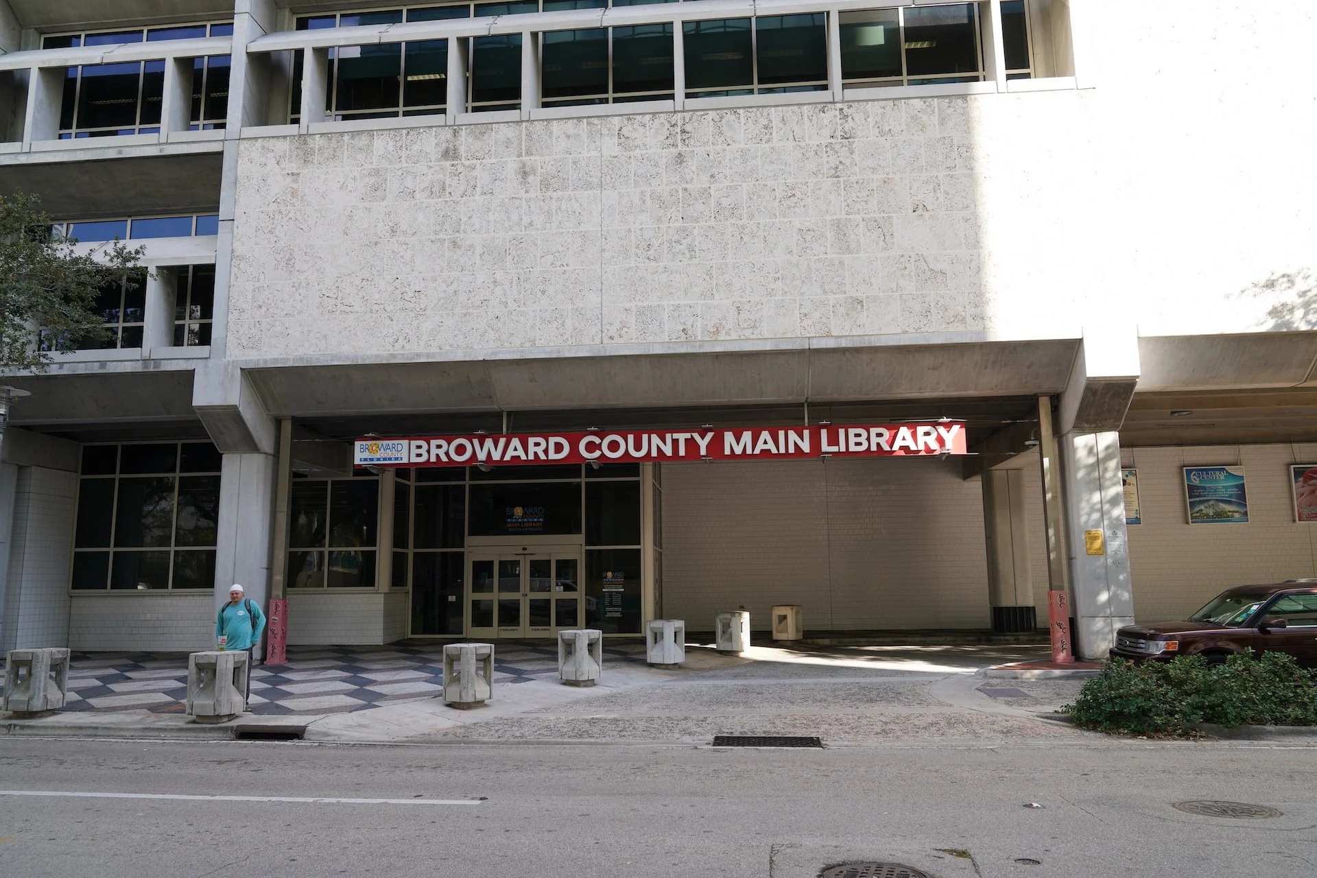 The Broward County Main Library building's entrance with a red and white sign displaying the library's name. There are large glass windows above the entrance, and a person is standing on the sidewalk near some barriers. A car is parked to the right.
