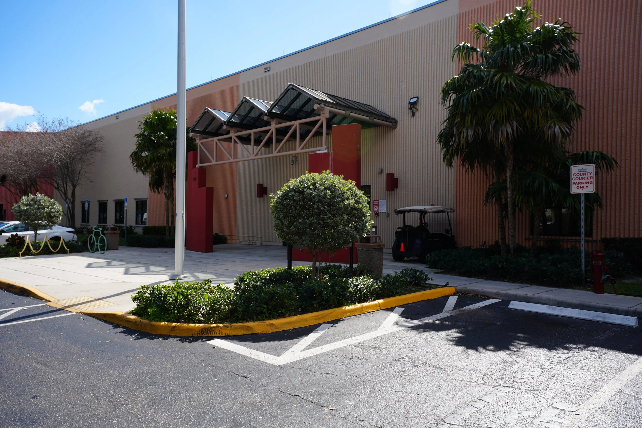 A parking lot in front of a large building with a textured beige and red exterior wall. There are trees, bushes, a golf cart, a sign indicating county courier parking, and a small covered area with solar panels.