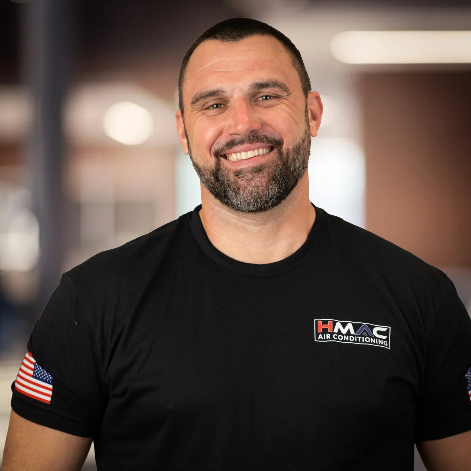 A man smiling, wearing a black T-shirt with an air conditioning company logo, standing indoors with blurred background.