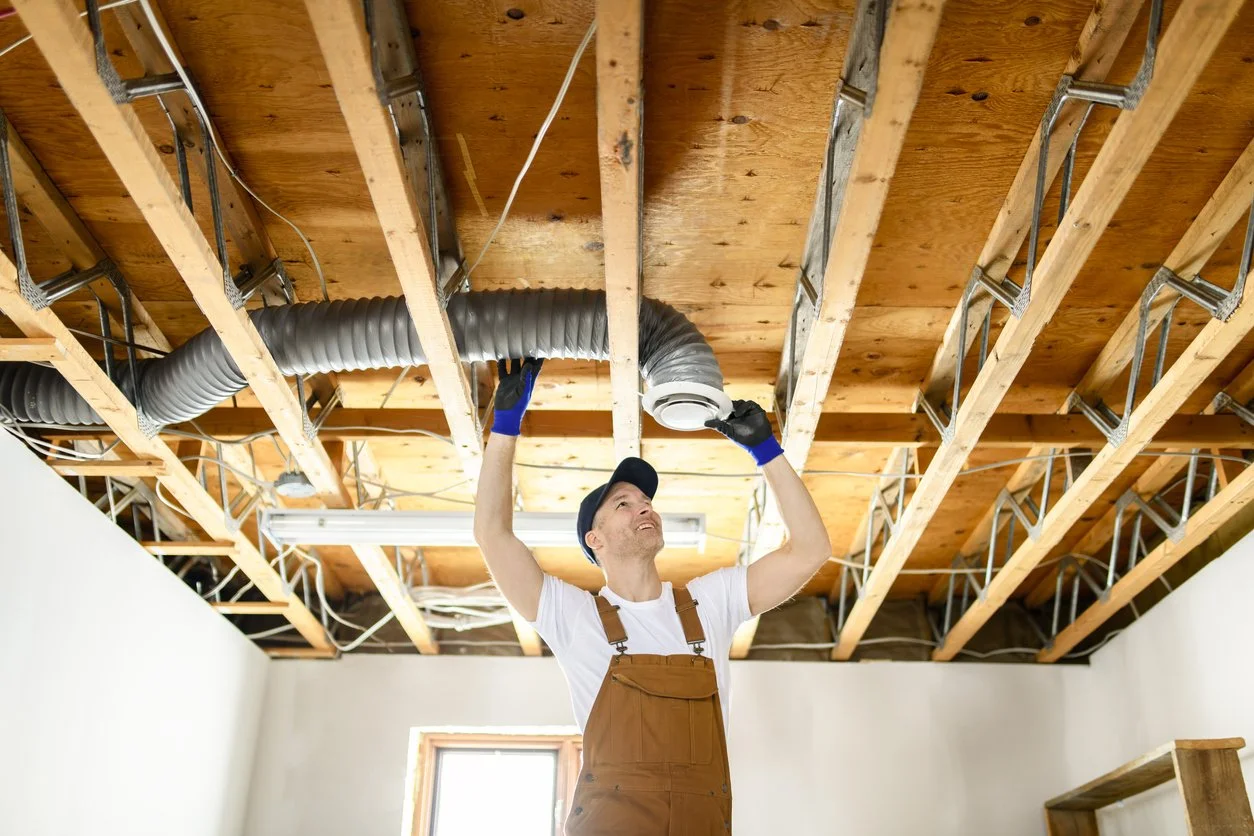 A man working on installing ductwork in a ceiling with exposed wood beams and electrical wiring.